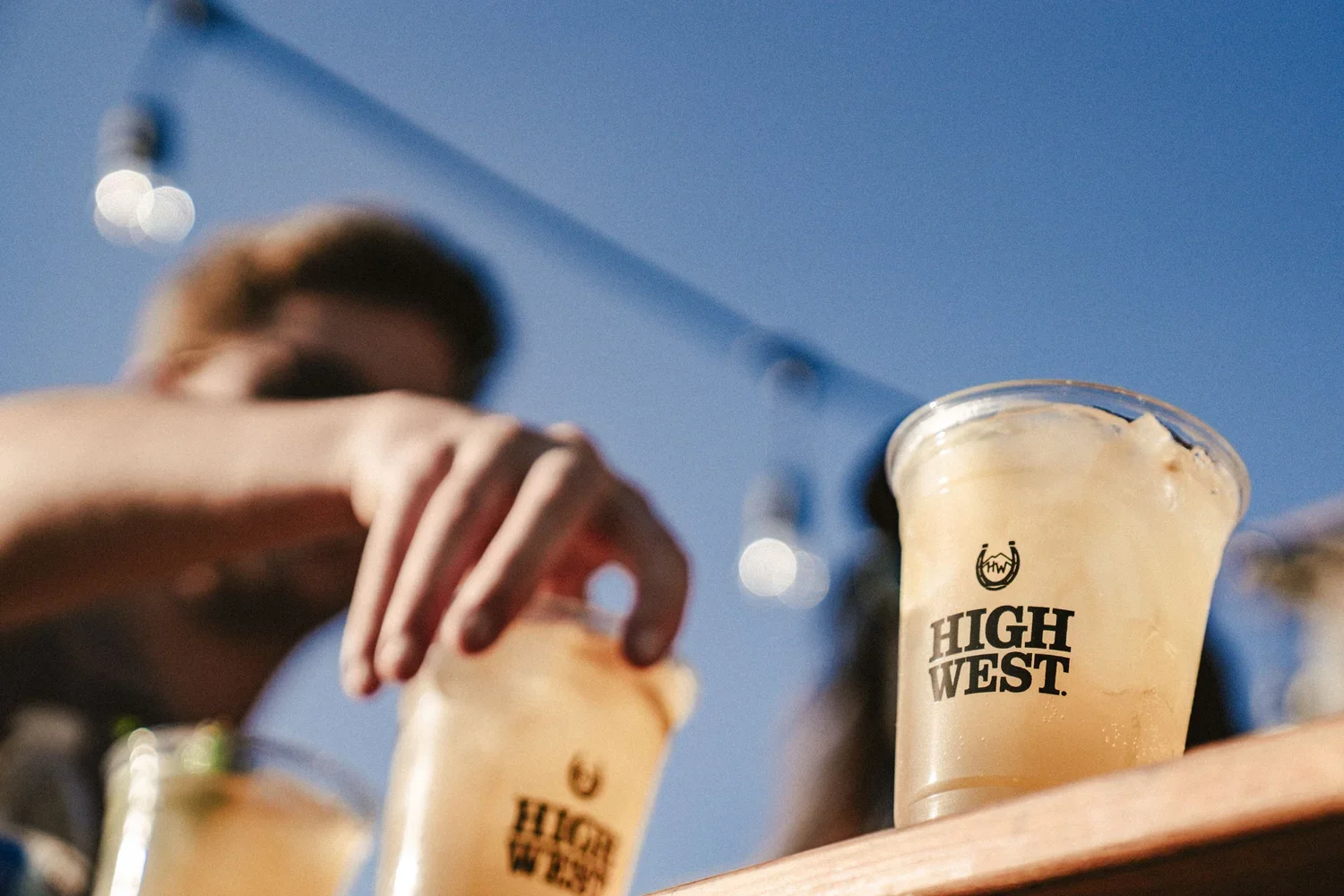 Person reaching for a plastic cup of a frozen Yellow High West cocktail on a wooden table outdoors, with a clear blue sky in the background.