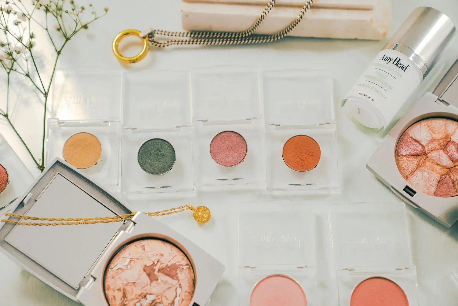 Makeup products on a white surface, including a marbled blush compact, four round pressed powders or eyeshadows in transparent cases, a green eyeshadow, a pink blush, a brownish-orange blush, and a beige blush. Also present are a white bottle labeled