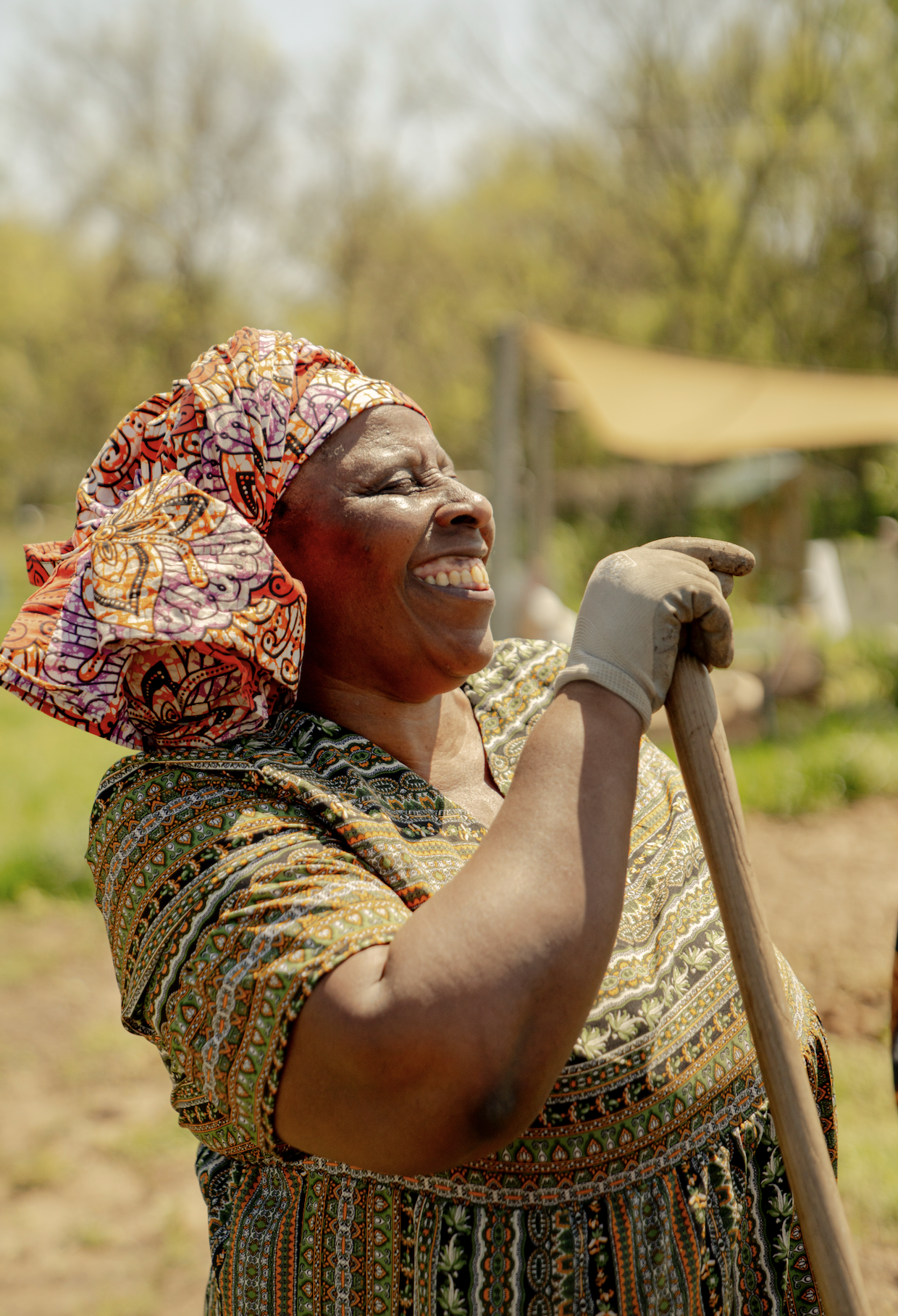 A joyful African woman wearing a colorful patterned headwrap and dress, holding a gardening tool outdoors with trees and a shade sail in the background.