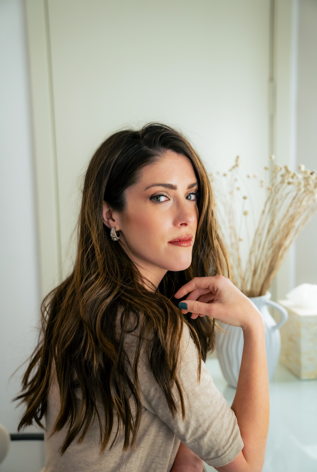 A woman with long wavy brown hair and blue eyes, wearing earrings and a beige top, sitting indoors with a neutral expression, hand near her chin, with dried plants and a pitcher in the background.
