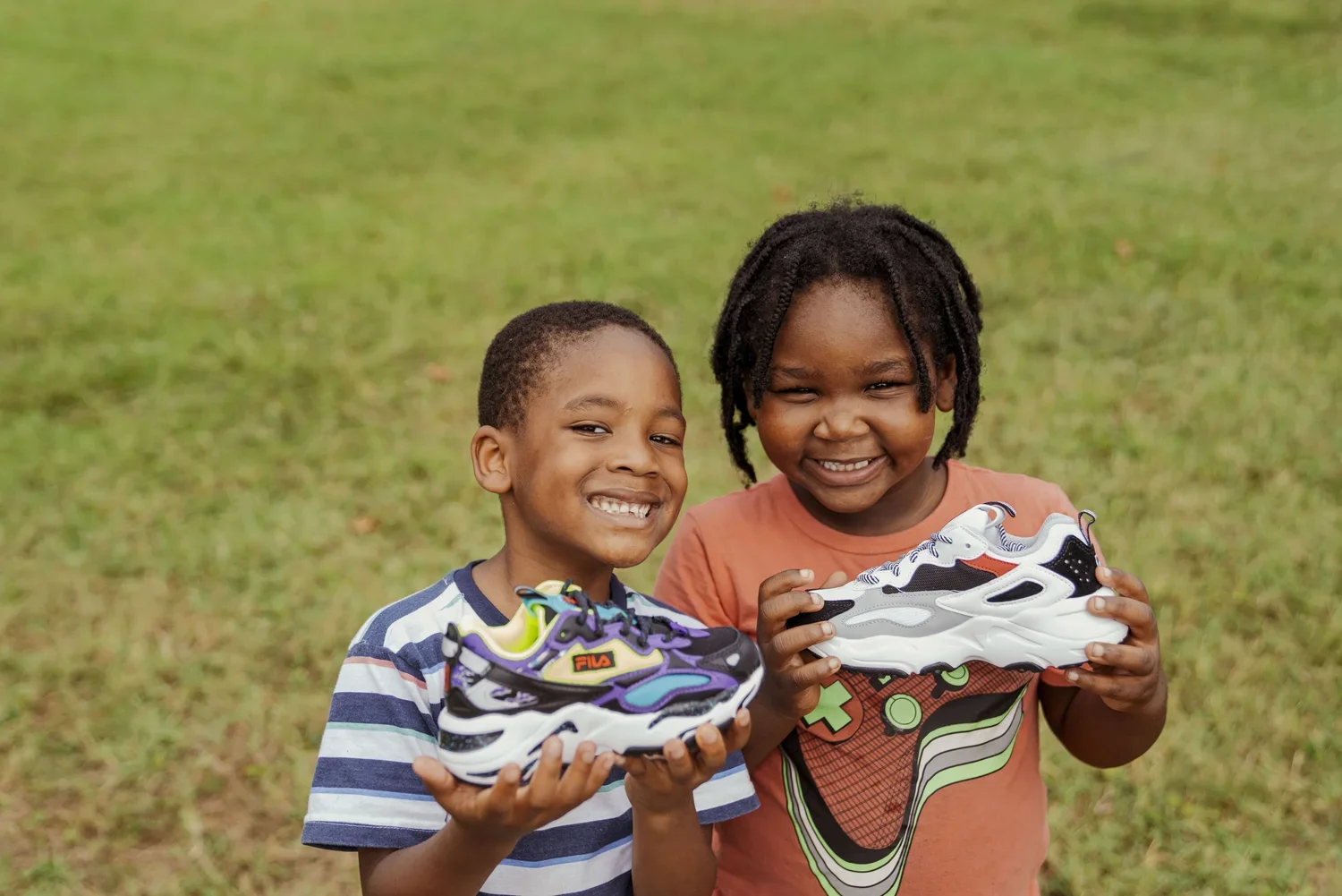 Two children smiling and holding colorful sneakers, standing outdoors on a grassy field.