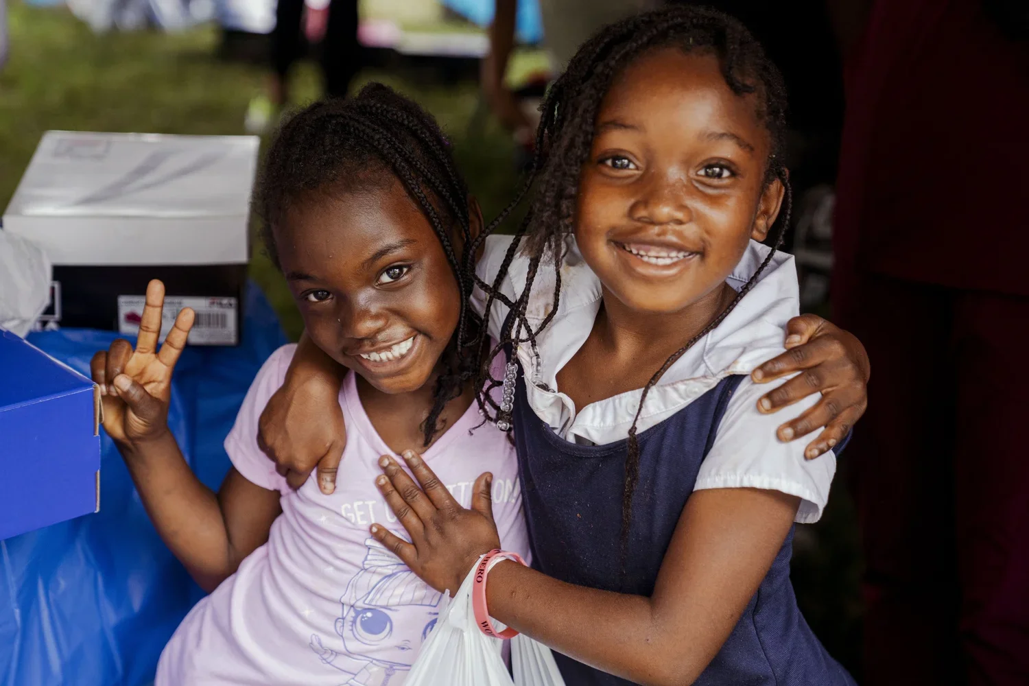Two young girls smiling and hugging each other outdoors. One girl is making a peace sign with her fingers.