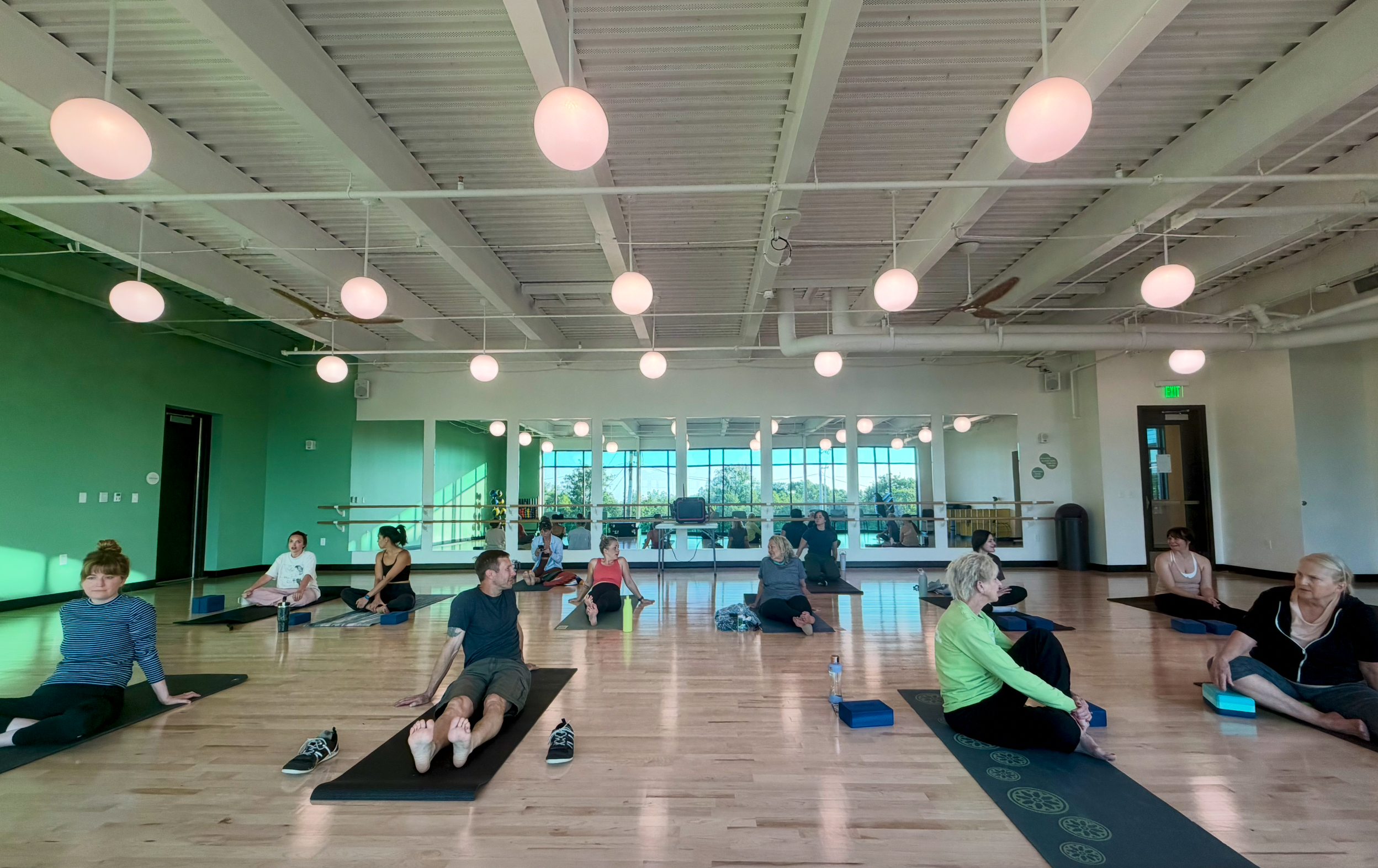 A group of people participating in a yoga or meditation class in a bright studio with large windows, white ceiling with hanging lights, and wooden floors.