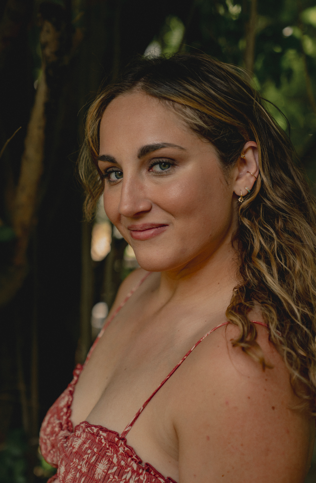 A woman with wavy hair and earrings, wearing a red floral spaghetti strap dress, standing outdoors among trees, facing slightly to the left, with a subtle smile.