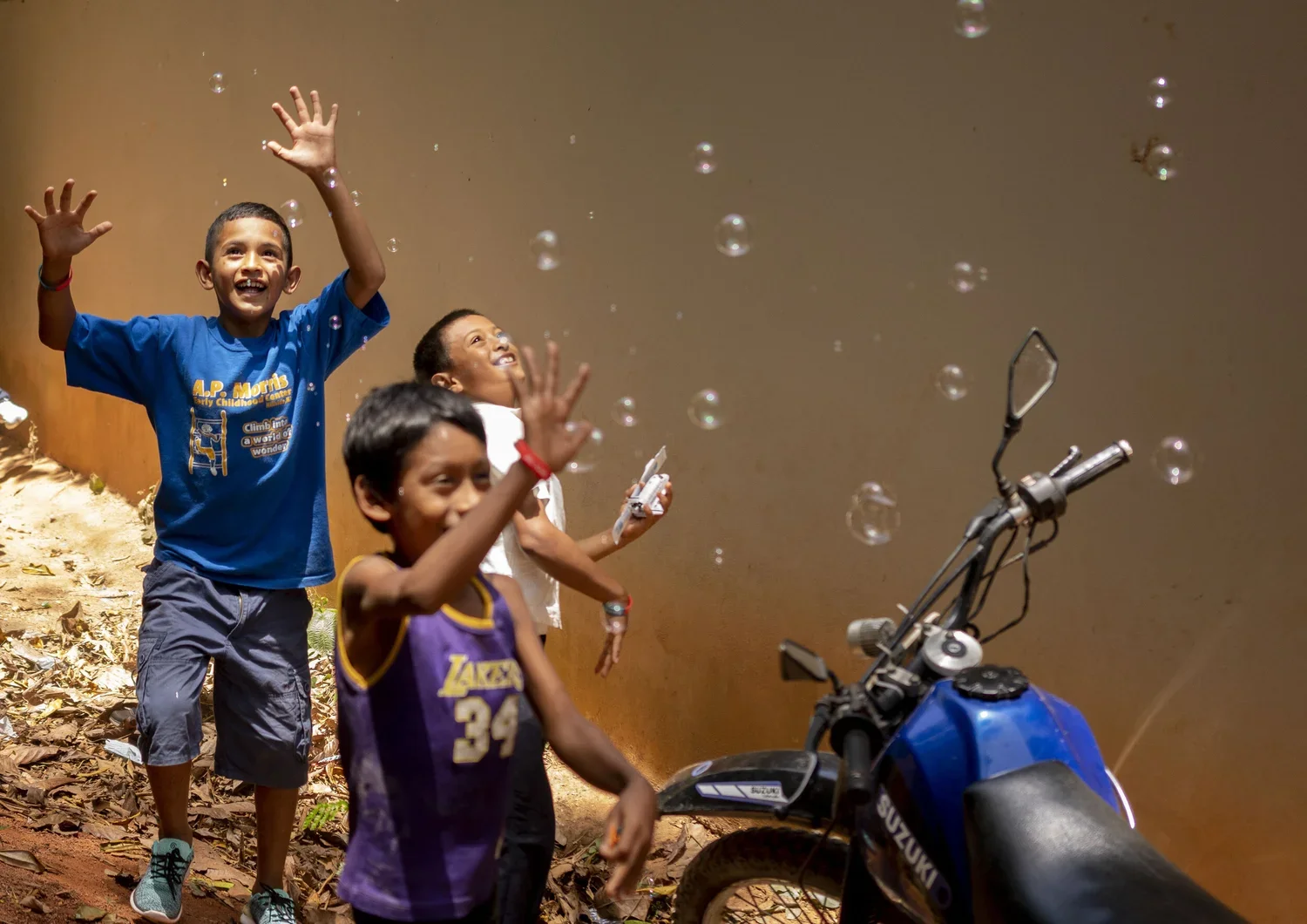 Three children are playing with soap bubbles near a motorcycle on a dirt path, with two of them reaching out towards the bubbles.
