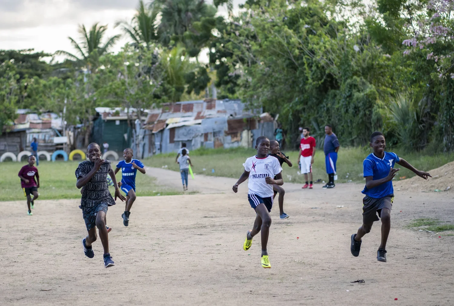 Children playing soccer on a dirt field in a rural area with makeshift houses and lush green trees in the background.