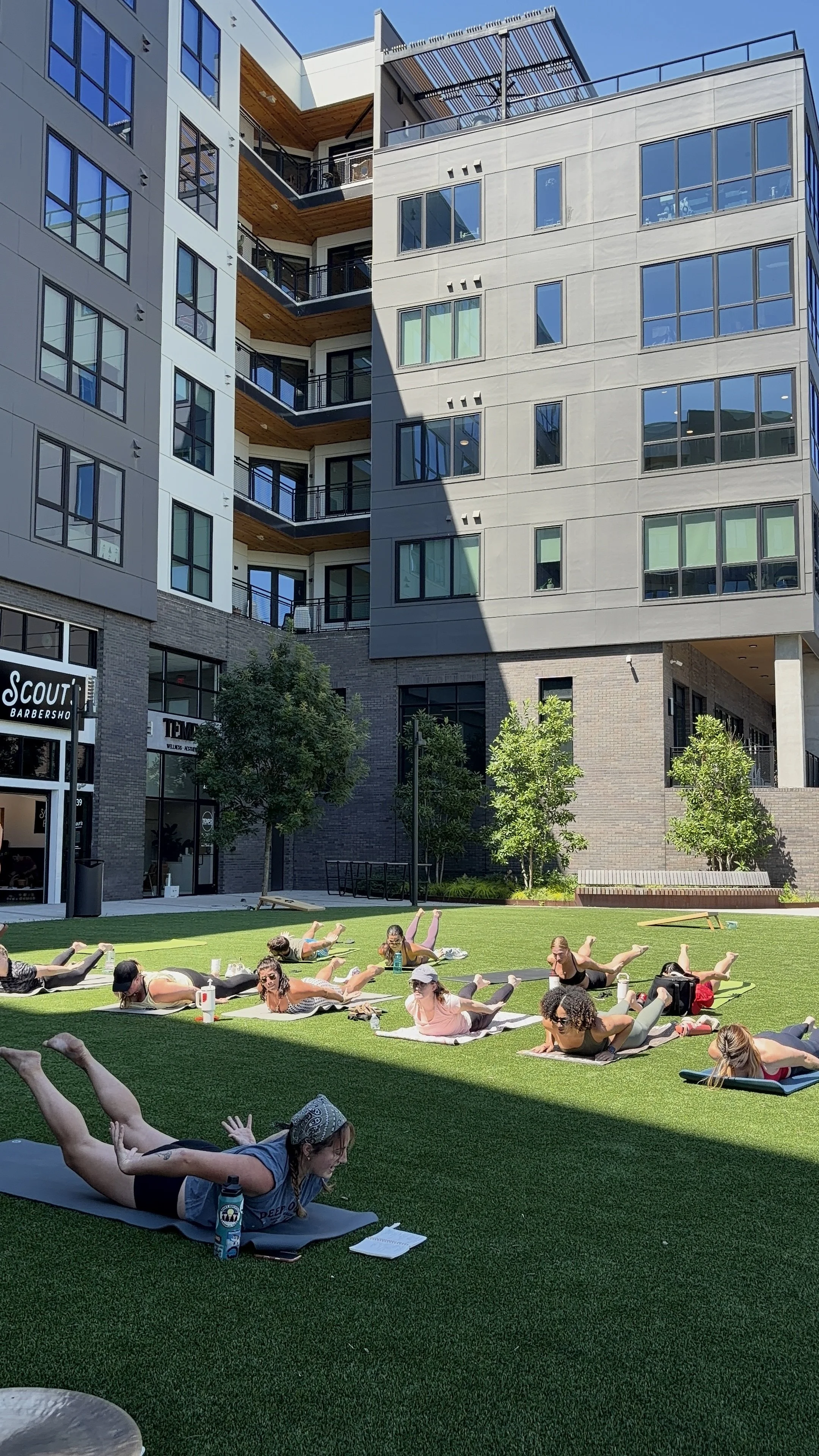 A group of people participating in an outdoor yoga class on a grassy area, with multi-story modern buildings in the background. Participants are lying on yoga mats, doing stretches and poses, with some reading or taking notes.