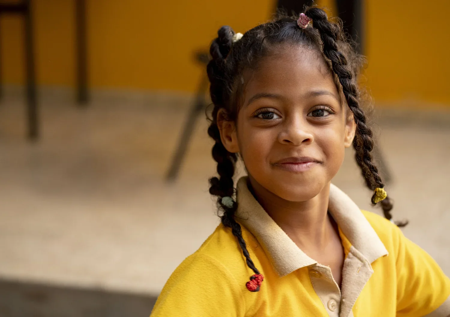 A young girl with braided hair adorned with colorful beads, smiling softly, wearing a yellow shirt, in a classroom with chairs and a yellow wall in the background.
