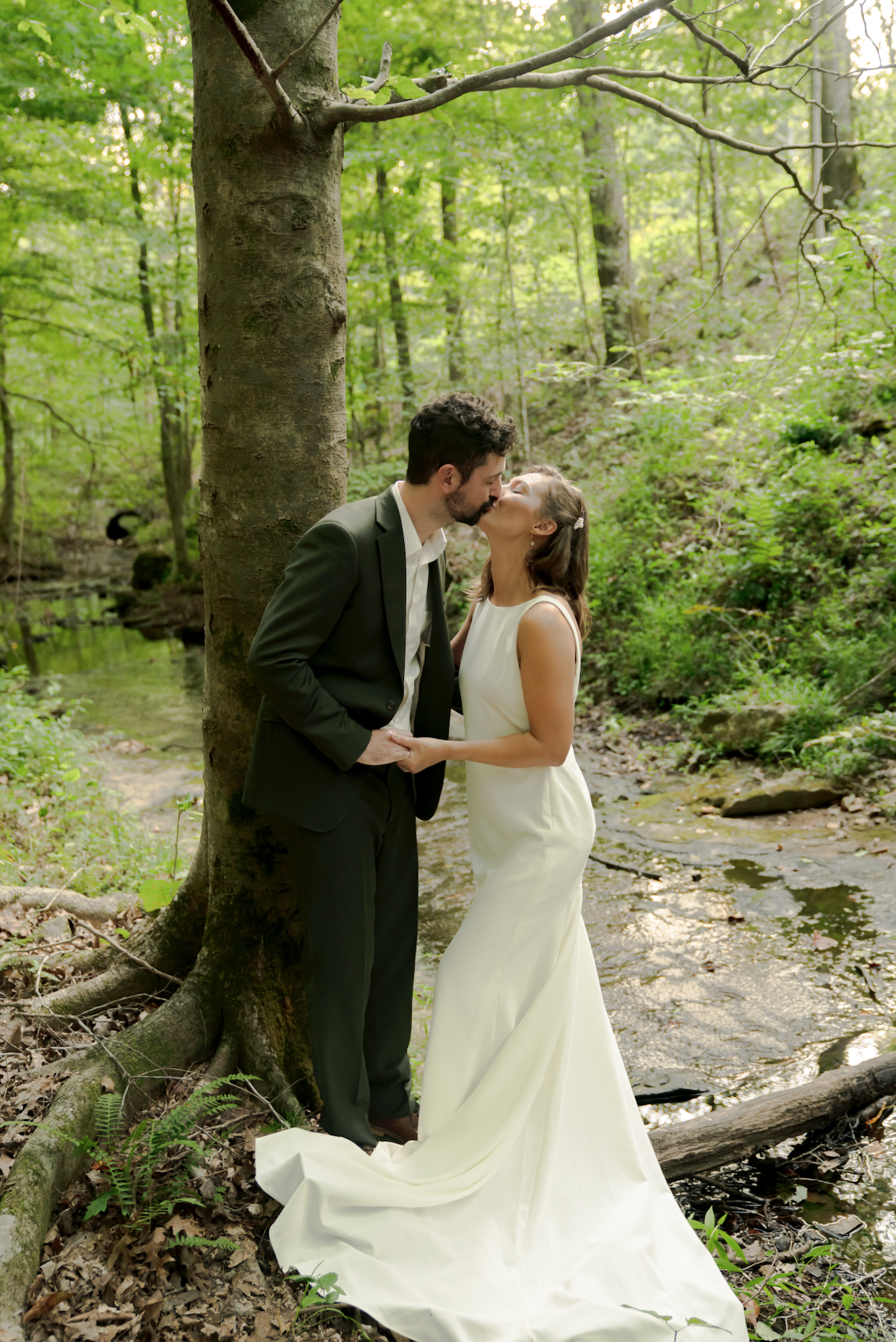 A bride and groom kiss in a green forest by a small creek, with the bride in a white dress and the groom in a dark suit.