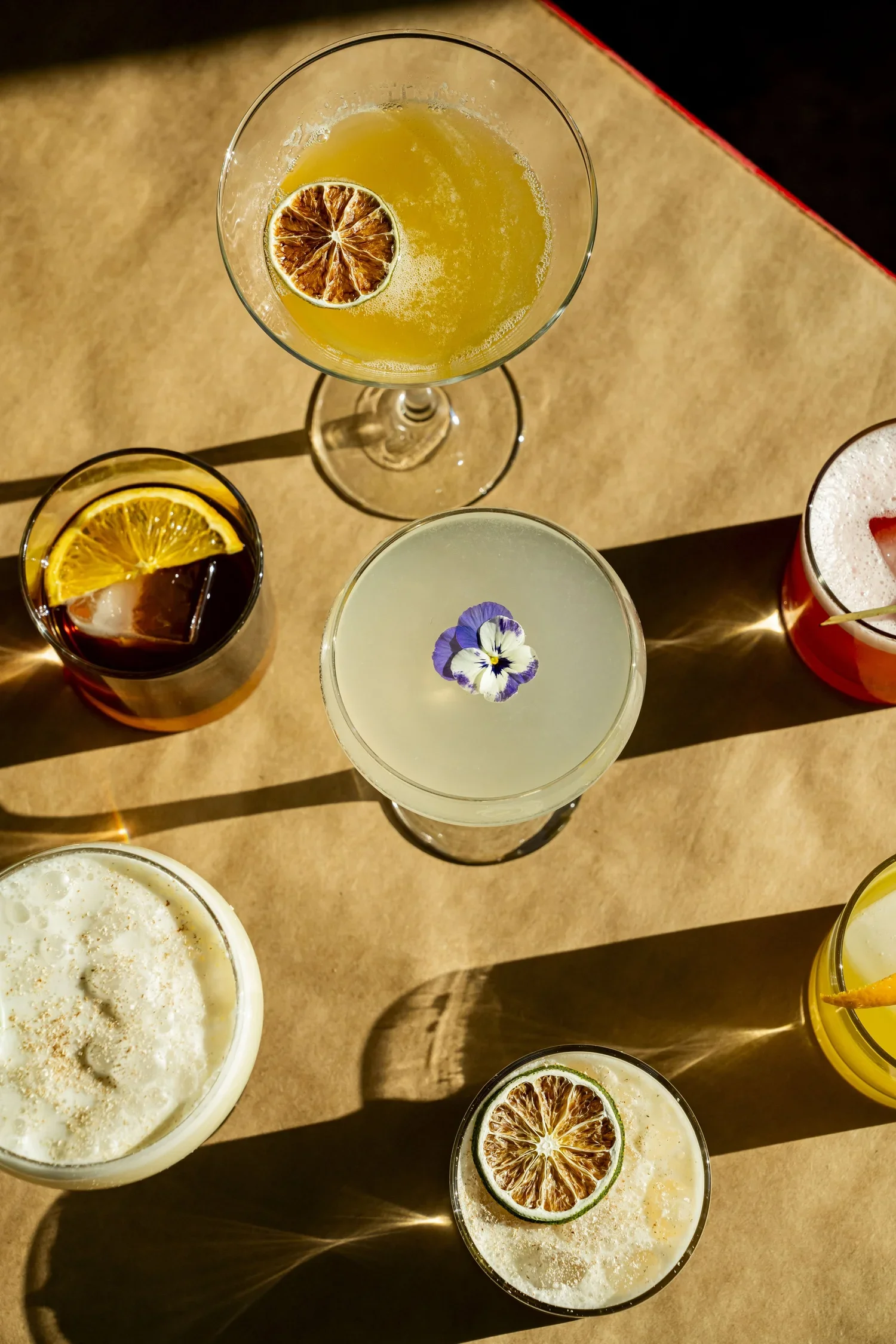 Various cocktails with fruit garnishes on a wooden table, illuminated by warm lighting creating shadows.