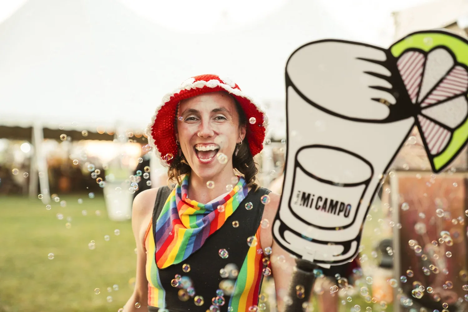 A woman smiling at a pride event, wearing a rainbow scarf and a red and white hat, holding a large sign with a graphic of a cocktail glass and the words 'El MICAMPO', with bubbles in the foreground.