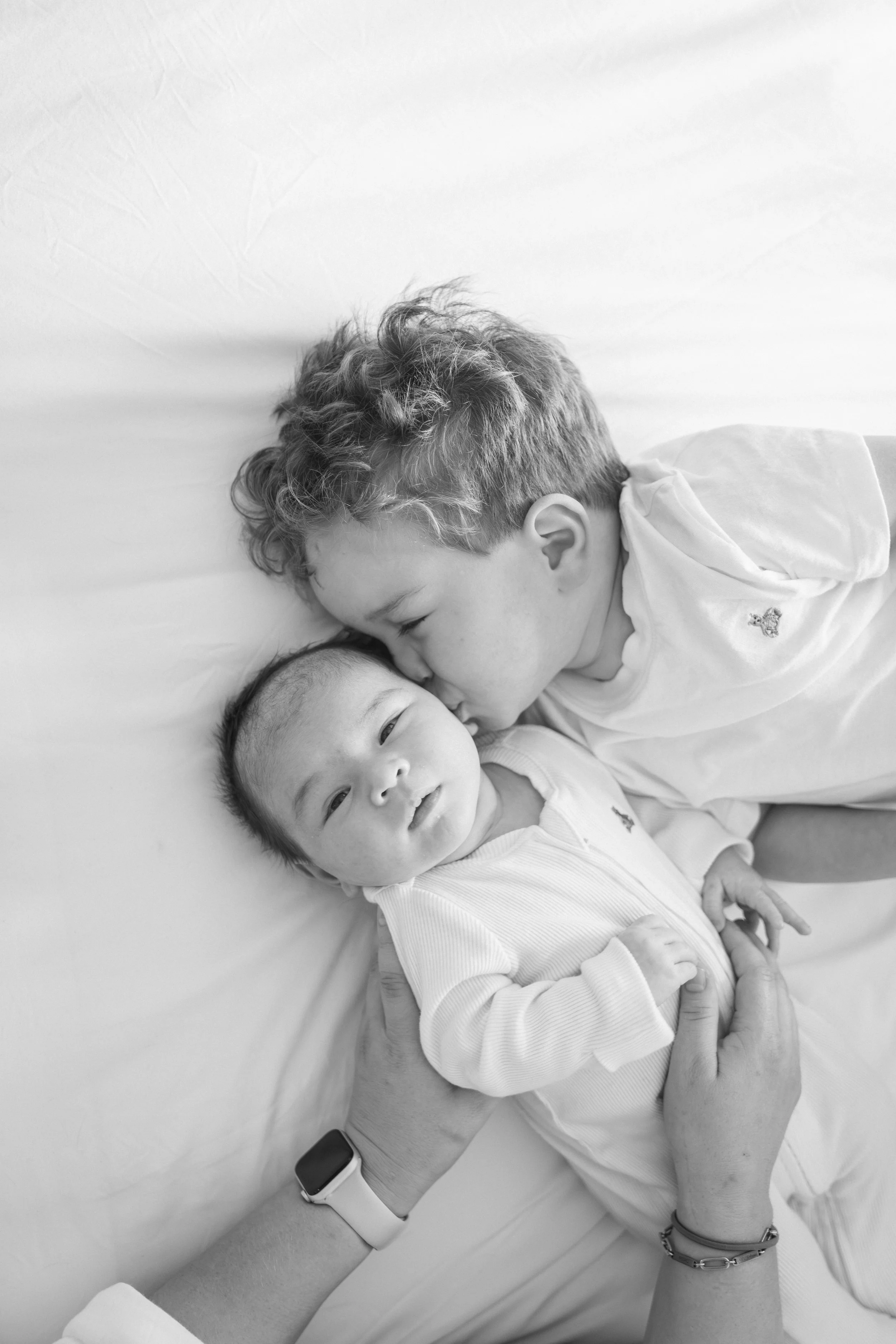 A young boy with curly hair is lying on a bed with eyes closed and a gentle smile, kissing a baby with short hair who is also lying on the bed, showing a loving interaction.