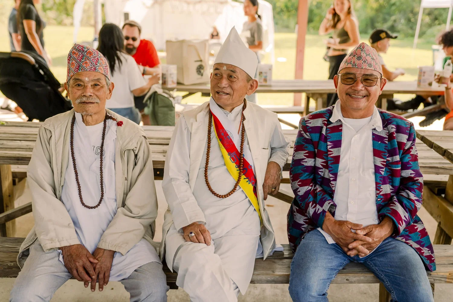 Three elderly men sitting on a wooden bench at an outdoor gathering, smiling. They are dressed in traditional attire and colorful clothing, with two wearing glasses and beaded necklaces. People are in the background, standing and socializing under a 