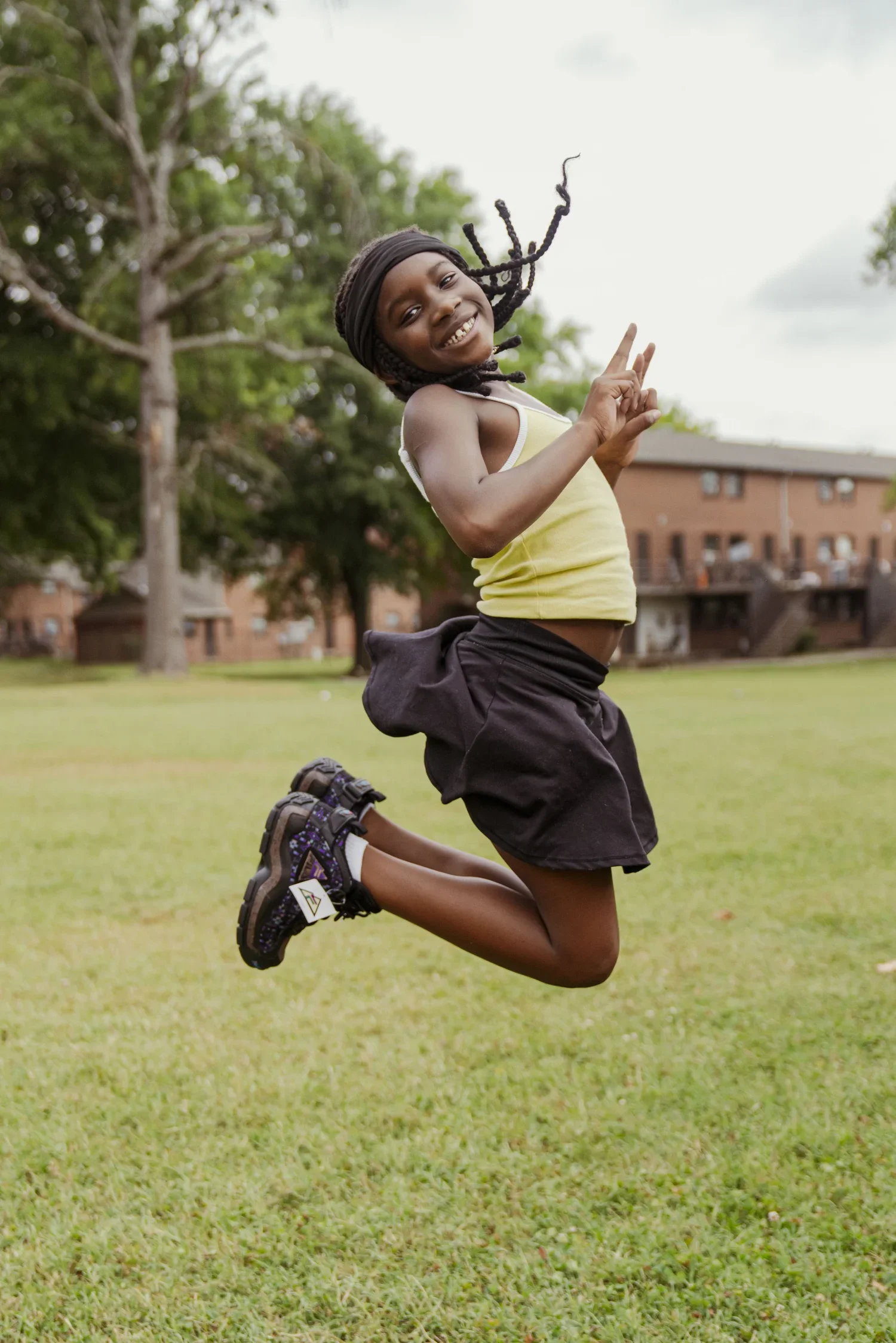 a young girl with a big smile, wearing a yellow tank top, black shorts, and sneakers, jumping in a grassy park with trees and brick buildings in the background, making a peace sign with her right hand.