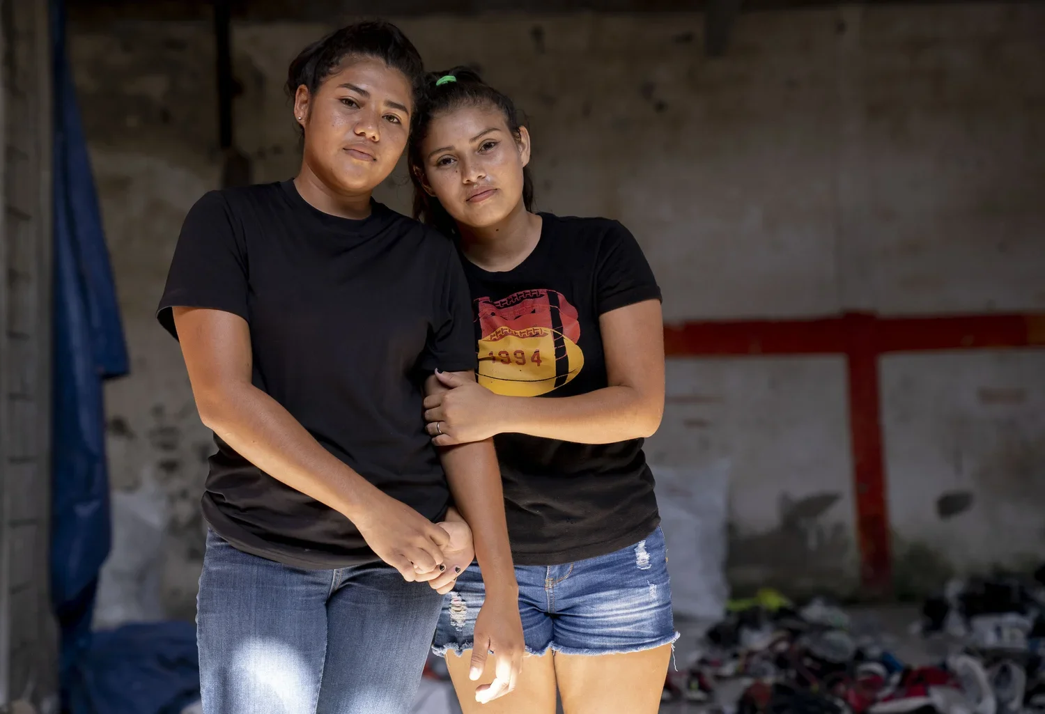 Two young women standing close together in an indoor setting with a rough wall background and scattered items on the floor, both wearing black t-shirts and denim shorts, one with her arm linked around the other's.