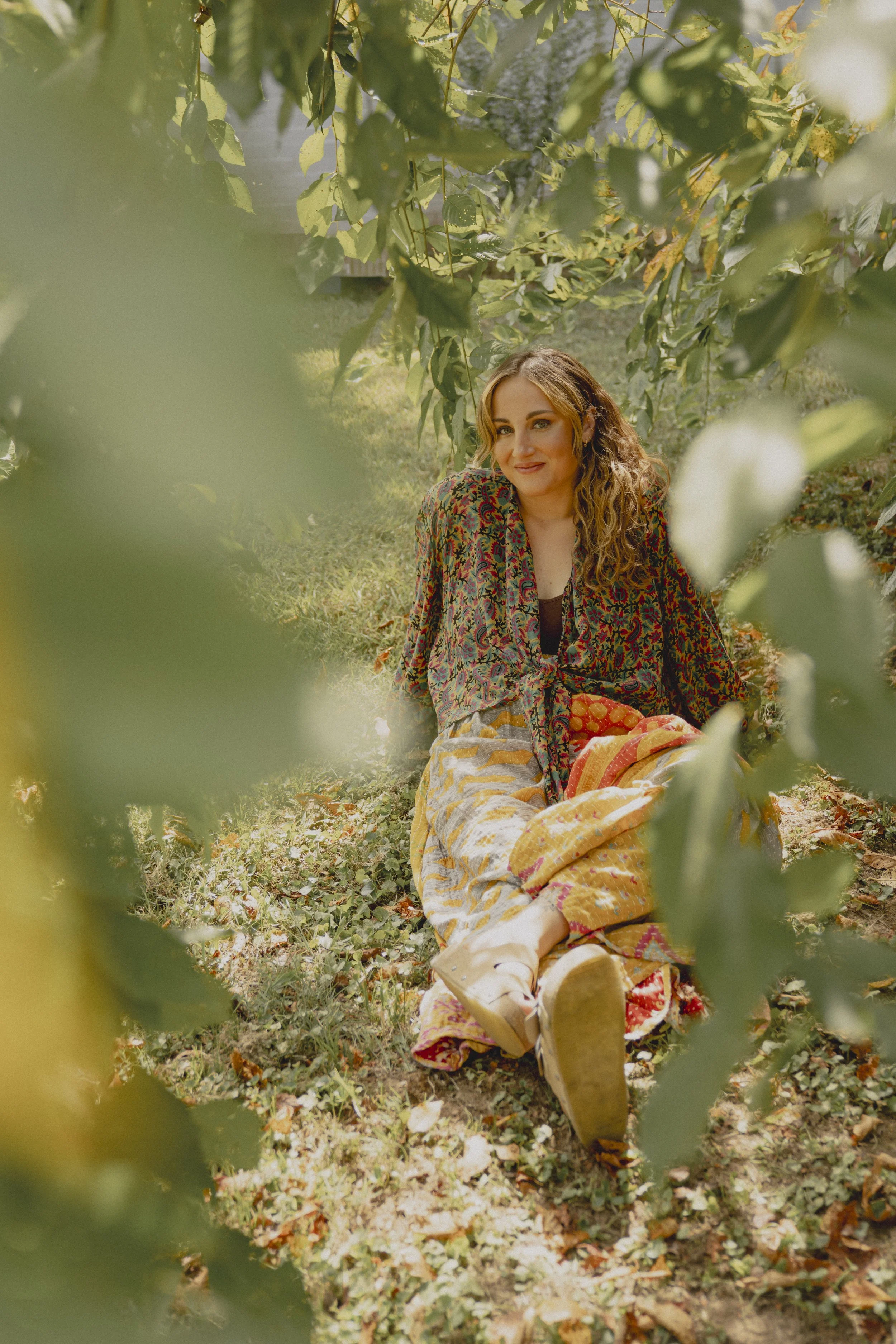 A woman sitting on the ground in an outdoor setting, partially obscured by green leaves in the foreground, with trees and sunlight in the background.
