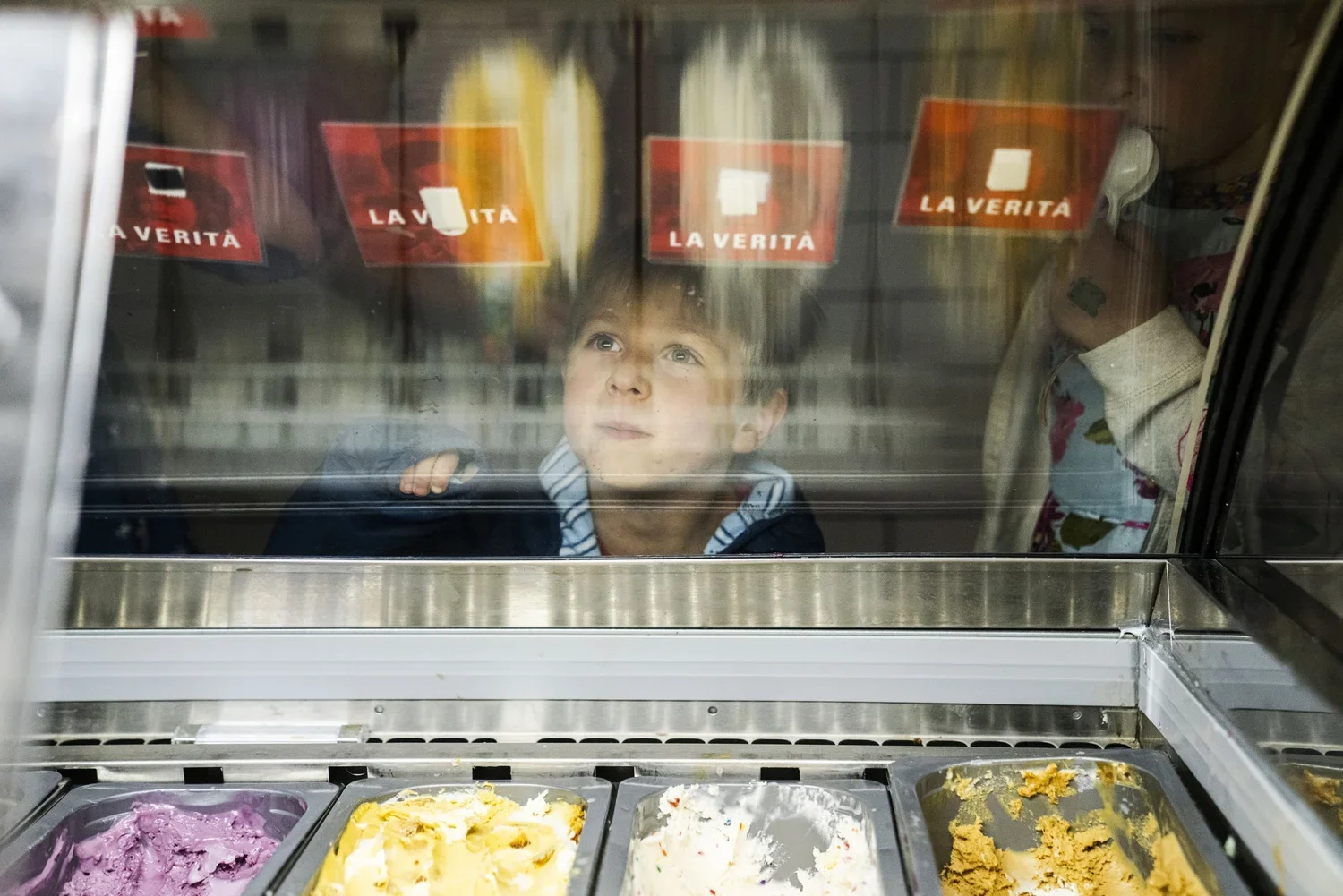 A young boy looks through the glass of an ice cream freezer at various flavors of ice cream inside, with red signs reading 'La Verità' on the glass.