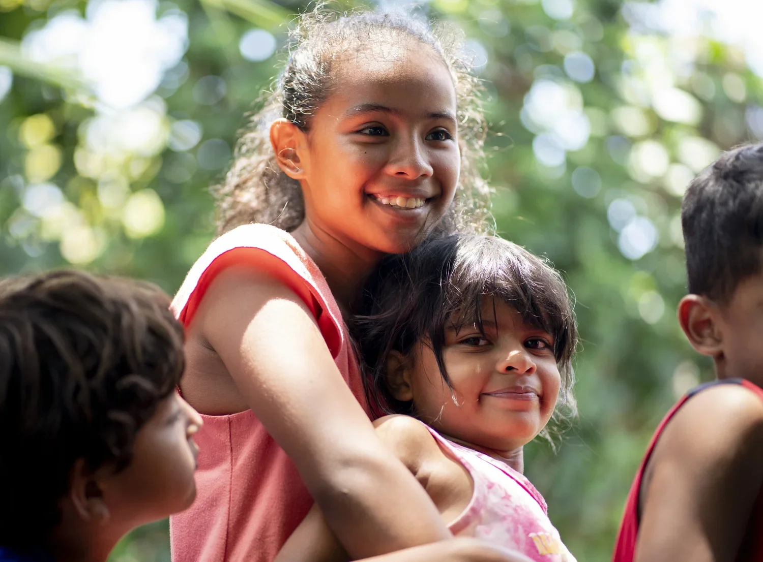 Smiling young girl with curly hair in a red dress sitting outdoors with children around her, green blurred background.