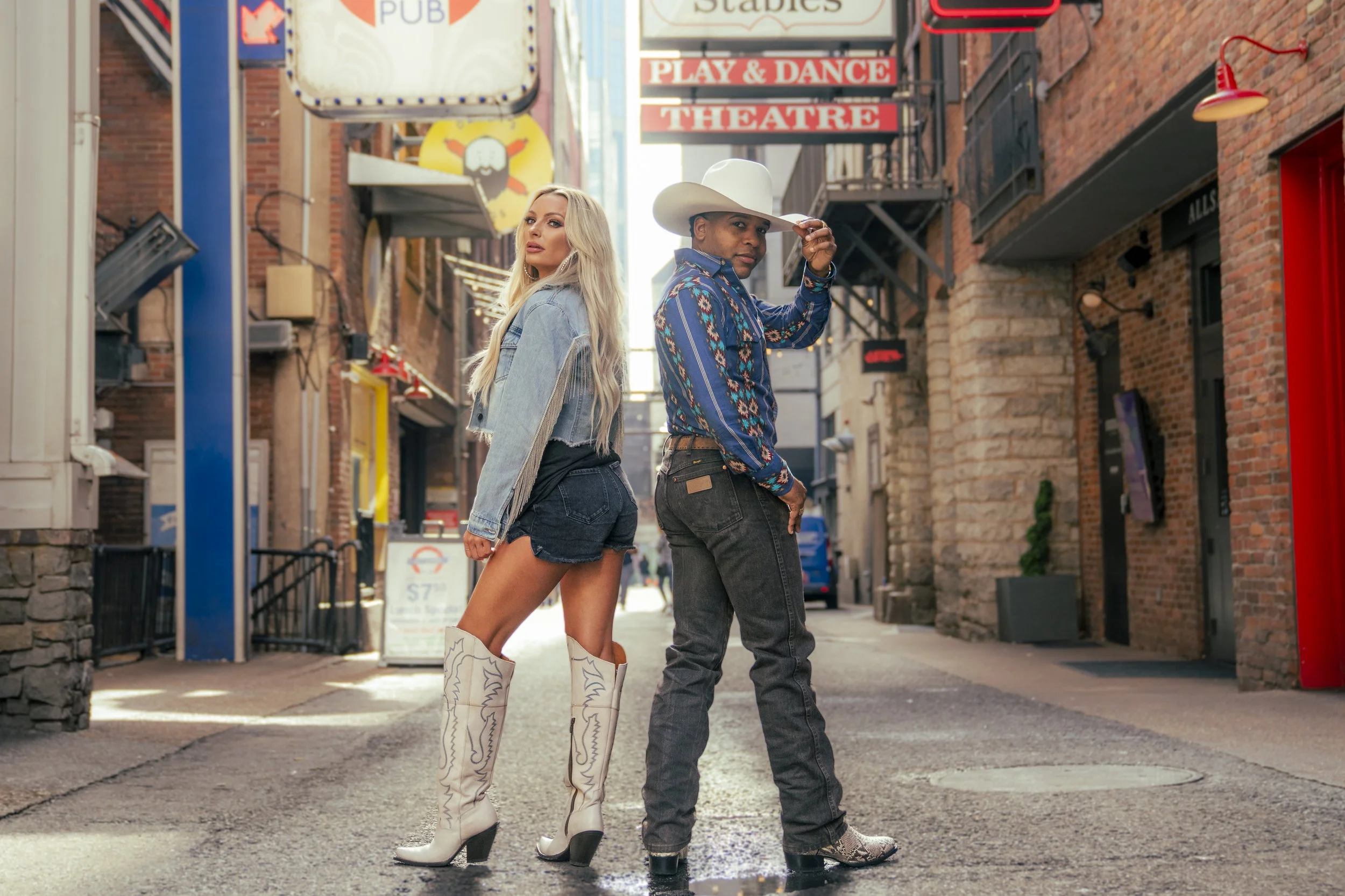 Two young adults, a woman with long blonde hair wearing a denim jacket, shorts, and white cowboy boots and a man wearing a large white cowboy hat, a blue patterned shirt, and dark jeans, are standing back-to-back on a city street, looking in opposite