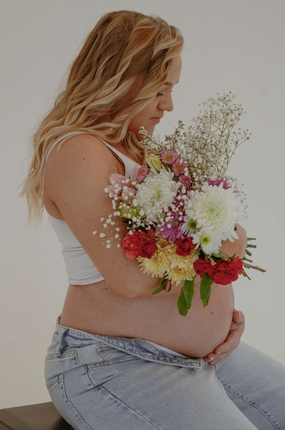 Pregnant woman with blonde wavy hair holding a bouquet of mixed flowers, wearing a white tank top and jeans, sitting against a plain white background.