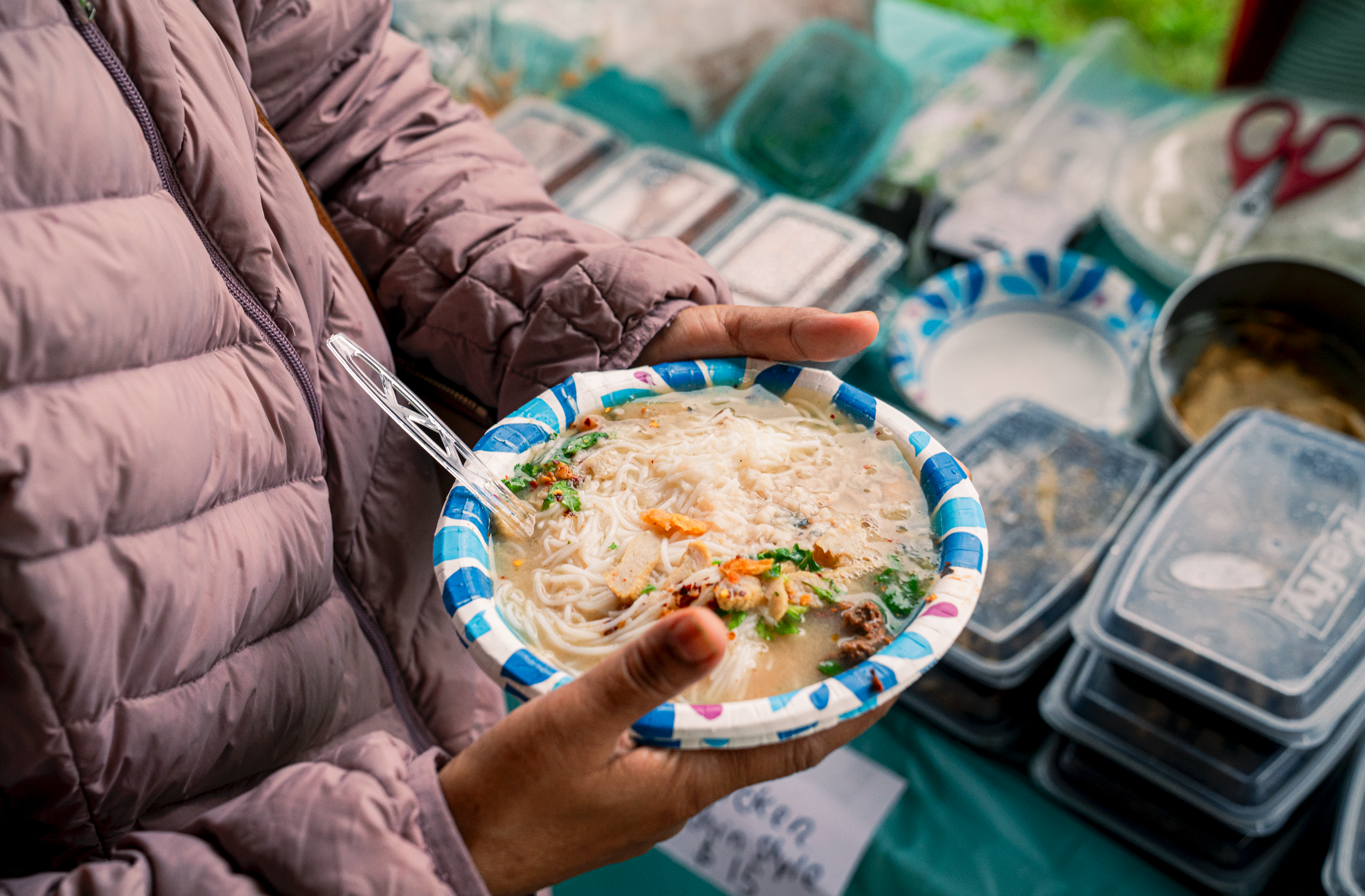 Person holding a bowl of melting cheese noodles with vegetables at a market stall, surrounded by various packaged food items and utensils.
