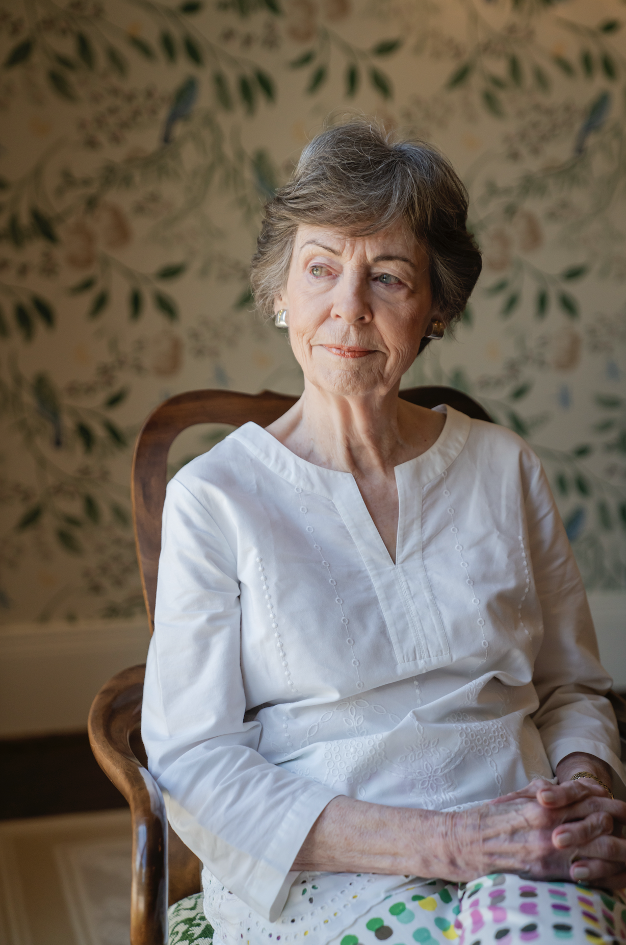 An elderly woman with short gray hair sitting in a wooden chair, wearing a white embroidered blouse, looking to her left with a thoughtful expression in front of a floral wallpaper background.