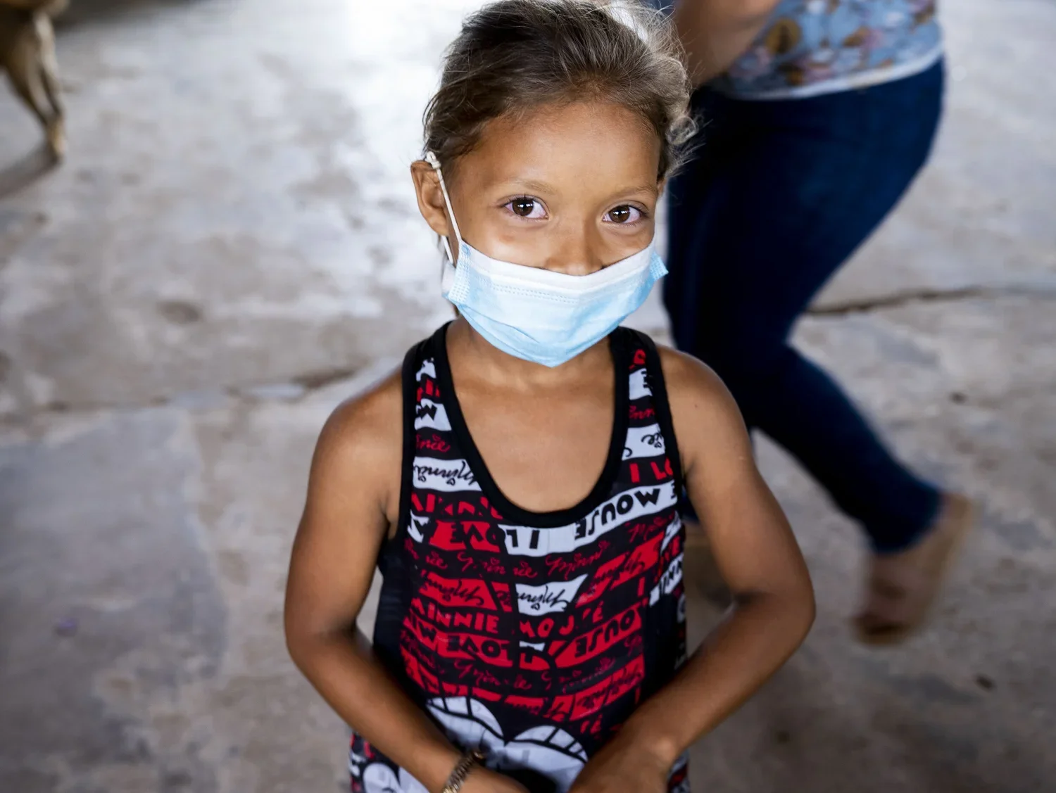 Young girl wearing a blue face mask and a black, red, and white patterned sleeveless dress, standing indoors on a concrete floor.