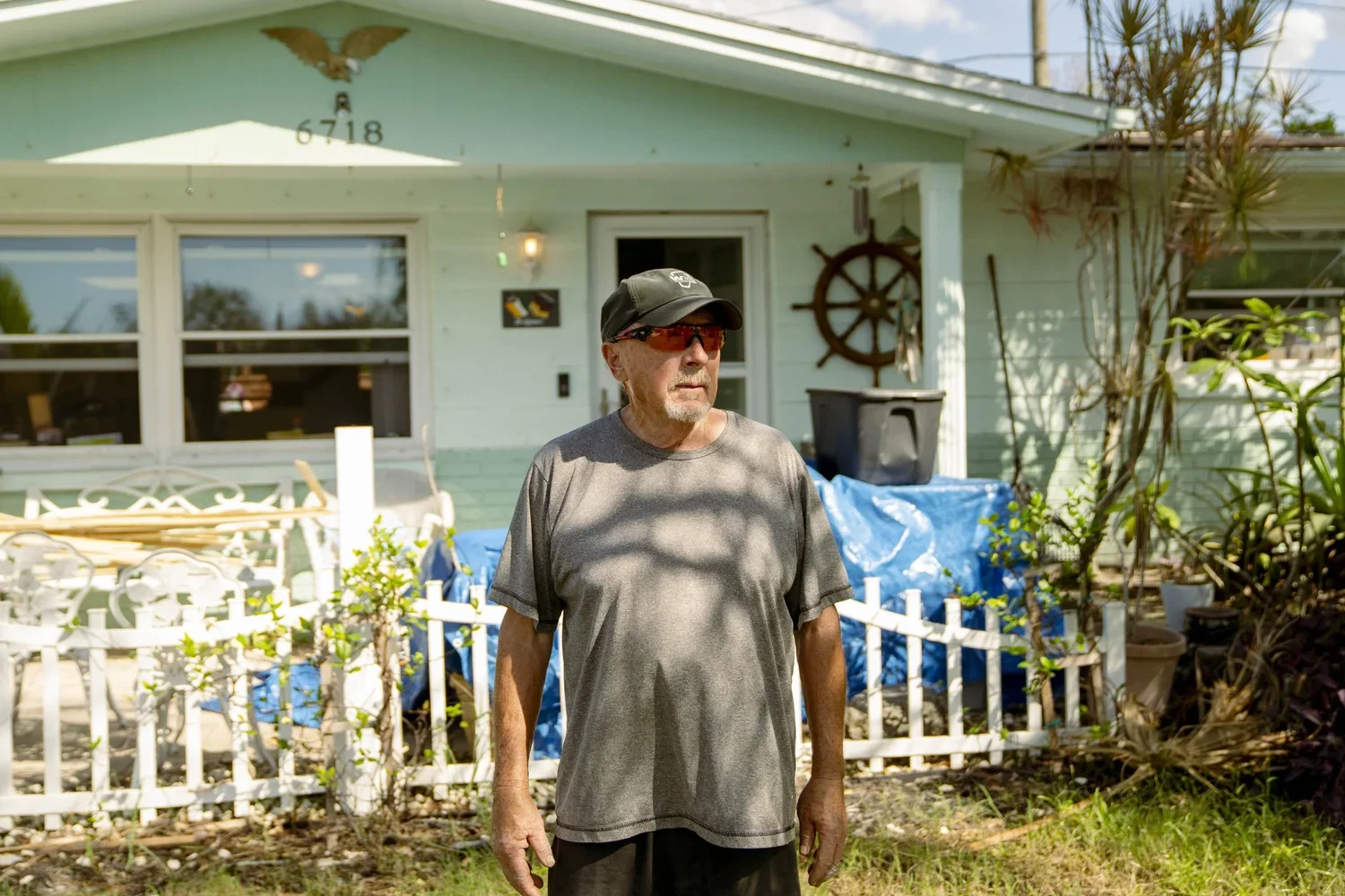 An older man with sunglasses and a cap stands outside a house with a light blue exterior. The house has a decorative wheel on the wall, and some plants and yard items are visible. The man is wearing a gray t-shirt and black shorts, and appears to be 