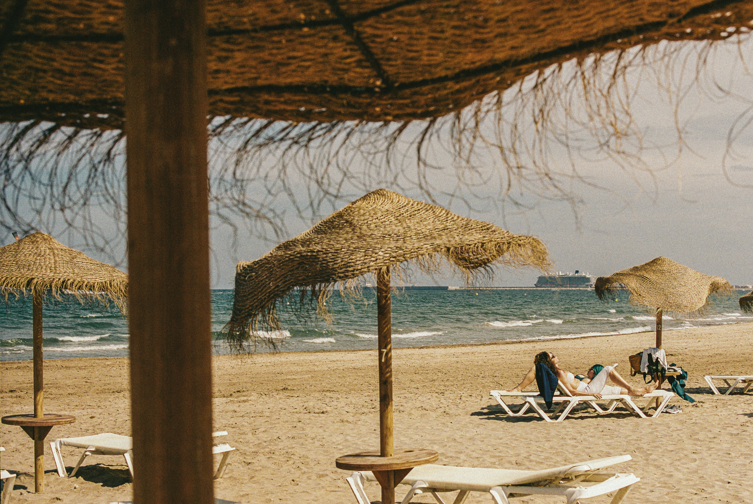 Beach scene with several straw umbrellas and lounge chairs along the sandy shore. A woman is sitting on a lounge chair under an umbrella near the water, with a bag nearby. In the background, there are cruise ships on the ocean, and a large ship is vi