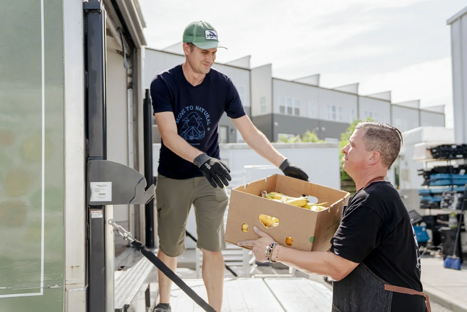 Two men exchanging a cardboard box of bananas outside a building, one man is on the truck unloading bananas, and the other man is receiving the box, both wearing casual clothing.