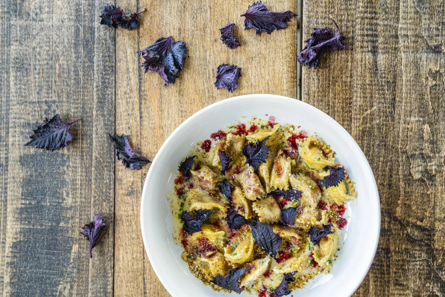 A bowl of cooked pasta with purple basil leaves on top, placed on a rustic wooden surface with scattered purple basil leaves around.
