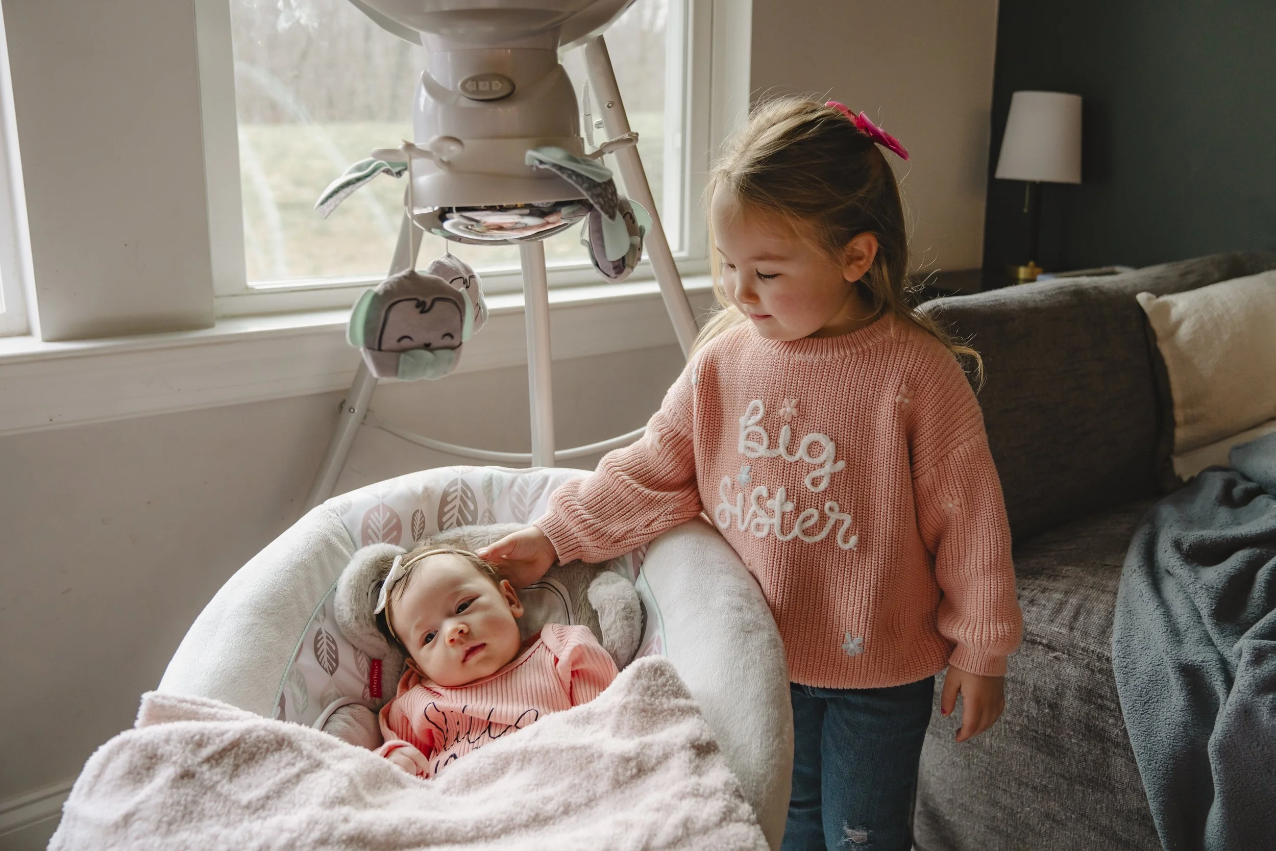 A young girl with a pink bow in her hair gently touches a baby lying in a bassinet. The baby is dressed in a pink sweater and surrounded by a soft blanket. The scene takes place near a window, with a dark-colored couch and a lamp in the background.