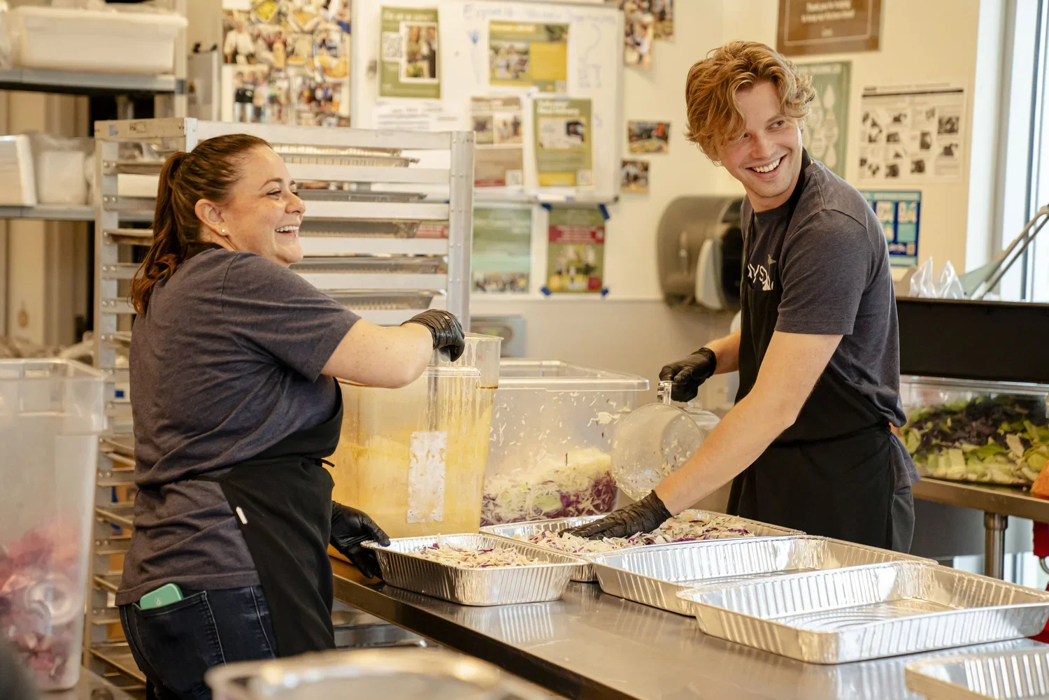 Two volunteers working together in a kitchen preparing food trays while smiling and wearing black gloves.