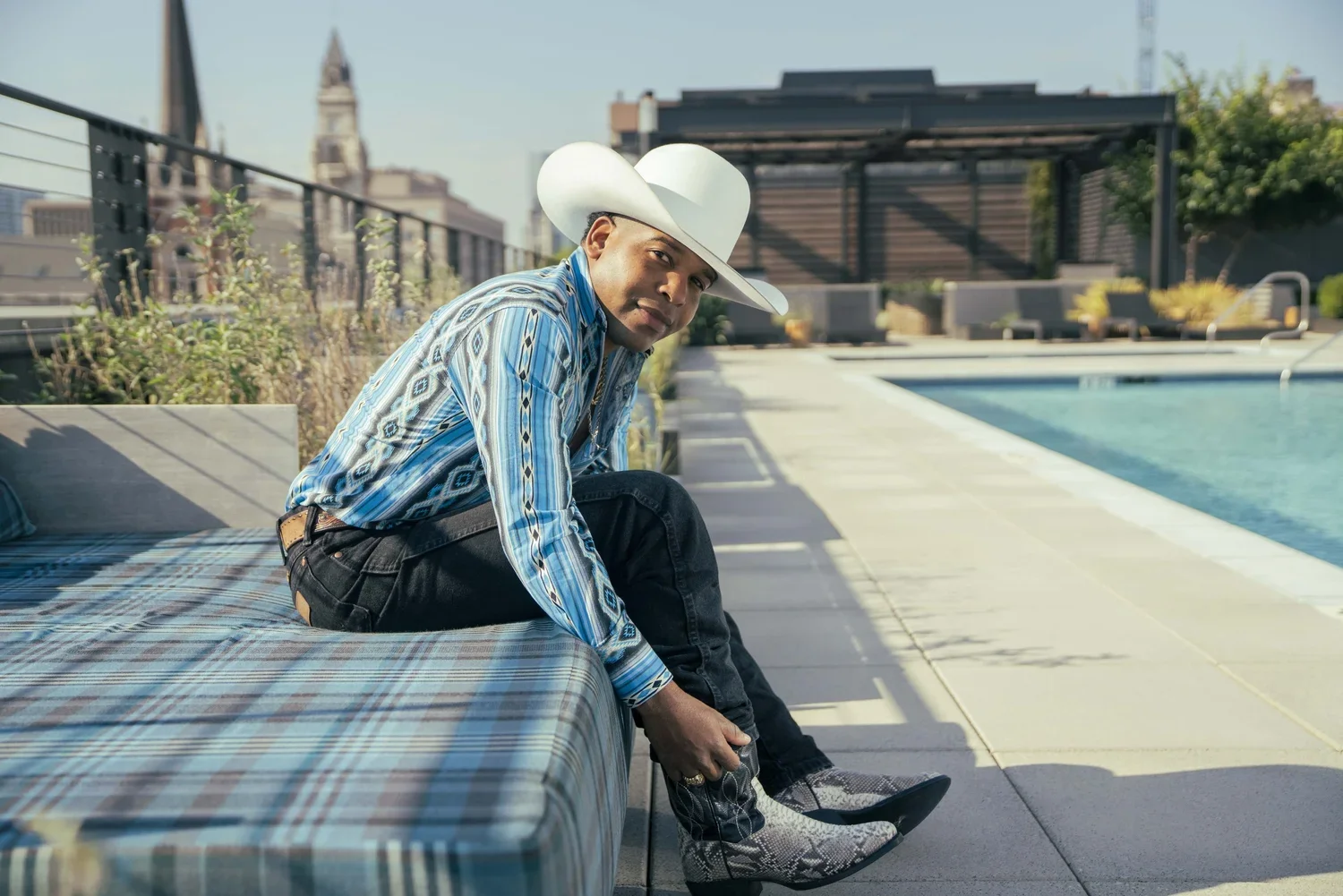 A man in a cowboy hat and patterned shirt sitting poolside, adjusting his boot.