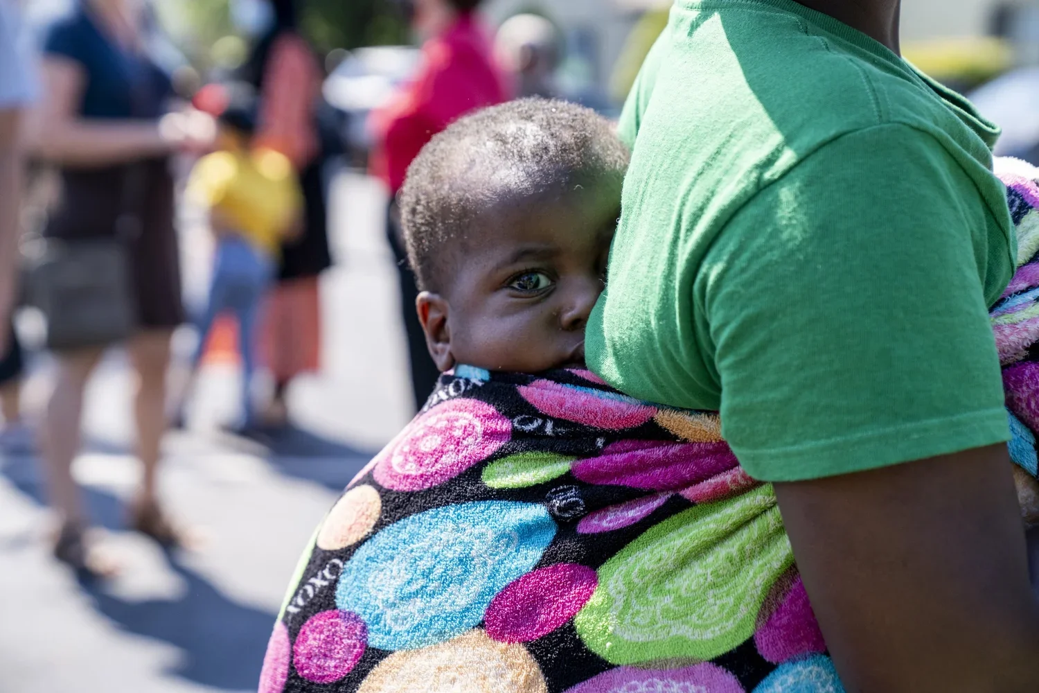 A young child with short hair is being carried on an adult's shoulder, wrapped in a colorful towel with circular patterns. The child is looking directly at the camera with a serious expression, while the adult is wearing a green shirt. People are blu