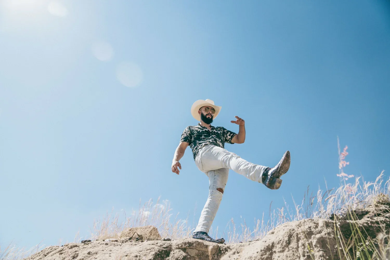 A man in a Hawaiian shirt, white jeans, sunglasses, and a cowboy hat balancing on one leg on a dirt hill under a clear blue sky.
