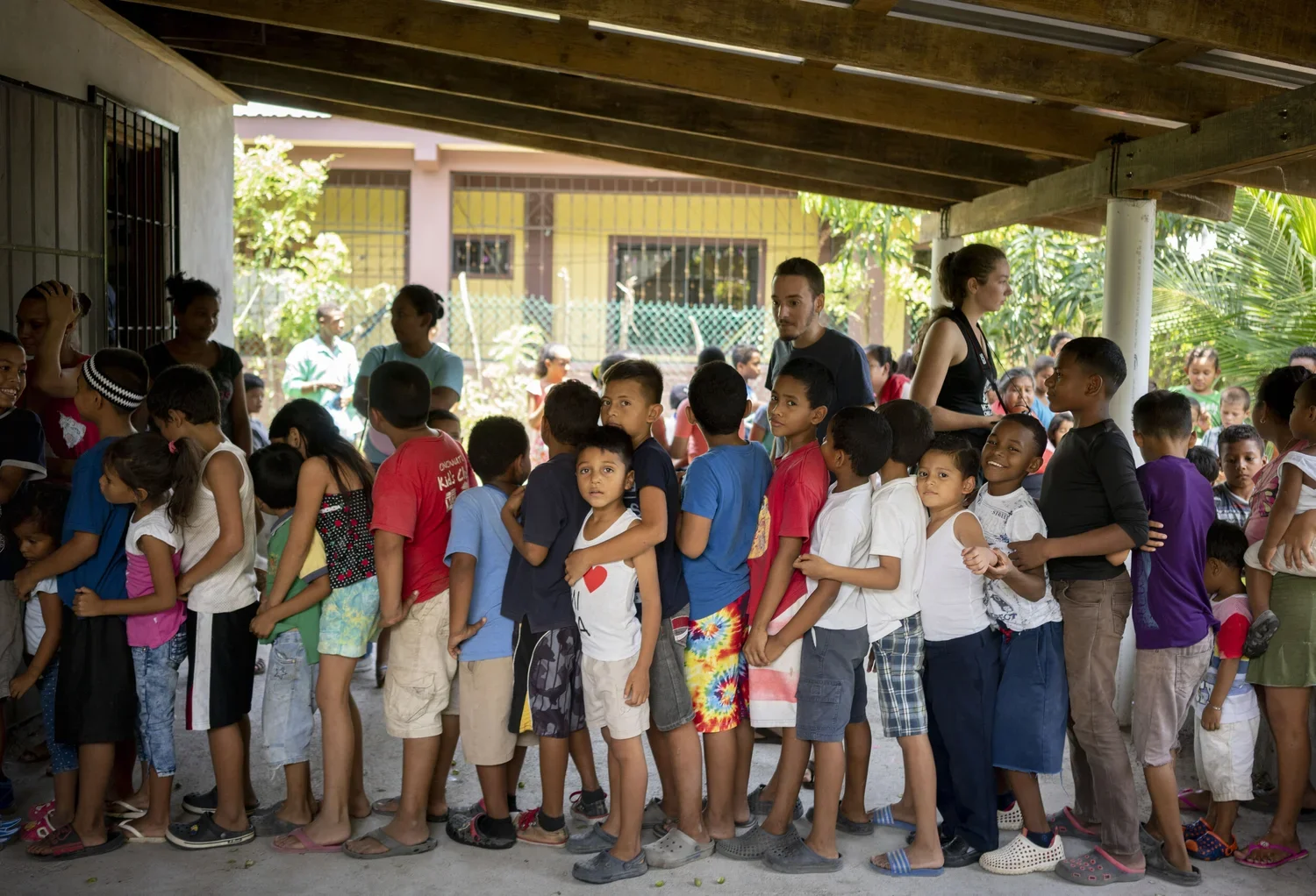 A group of children standing in a line outside of a building, with several adults supervising and chatting nearby.