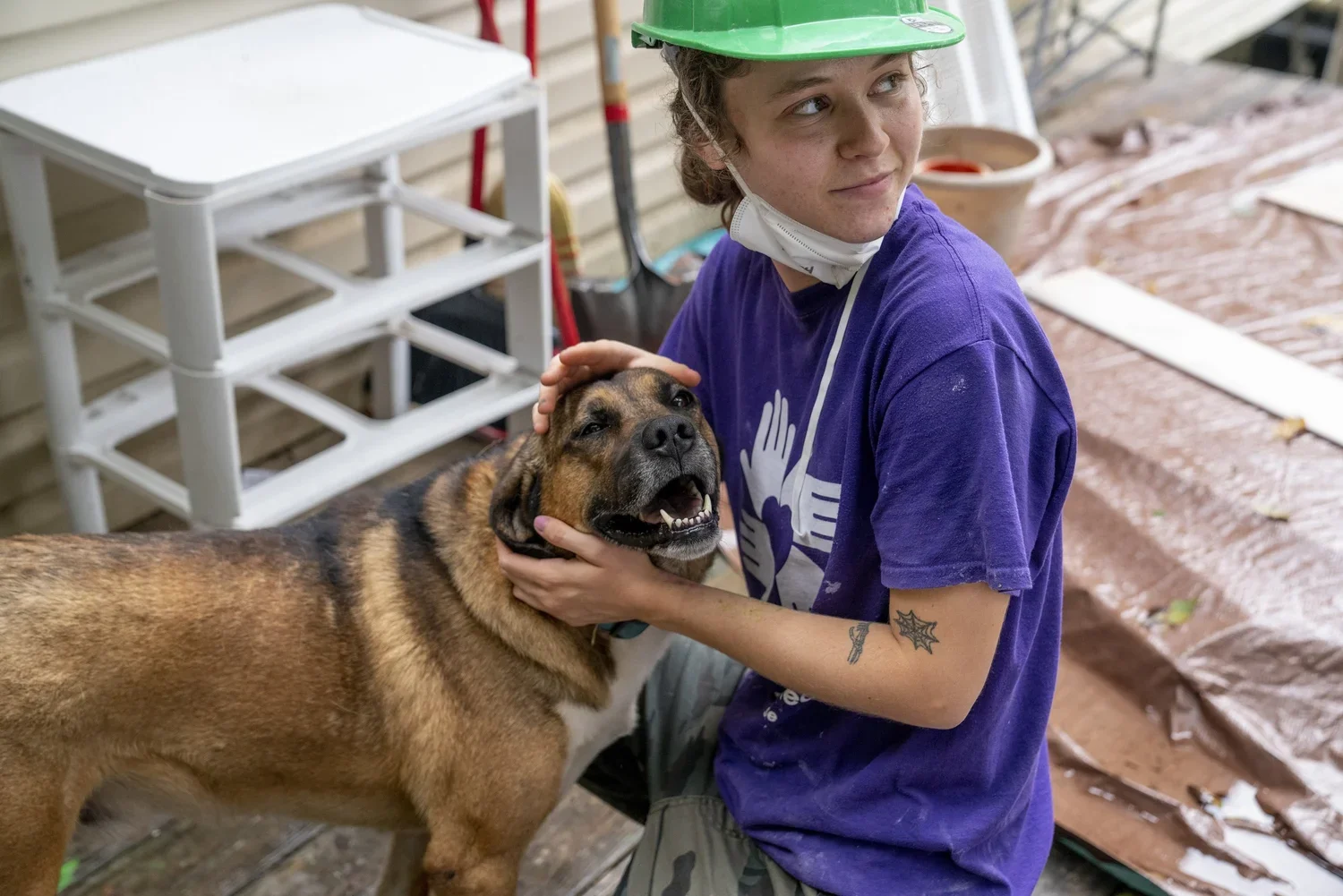 A young woman wearing a green safety helmet and a purple t-shirt is petting a large brown and black dog, which appears to be a boxer mix, outdoors near a wooden building and a white shelving unit. The woman is smiling and has tattoos on her right arm