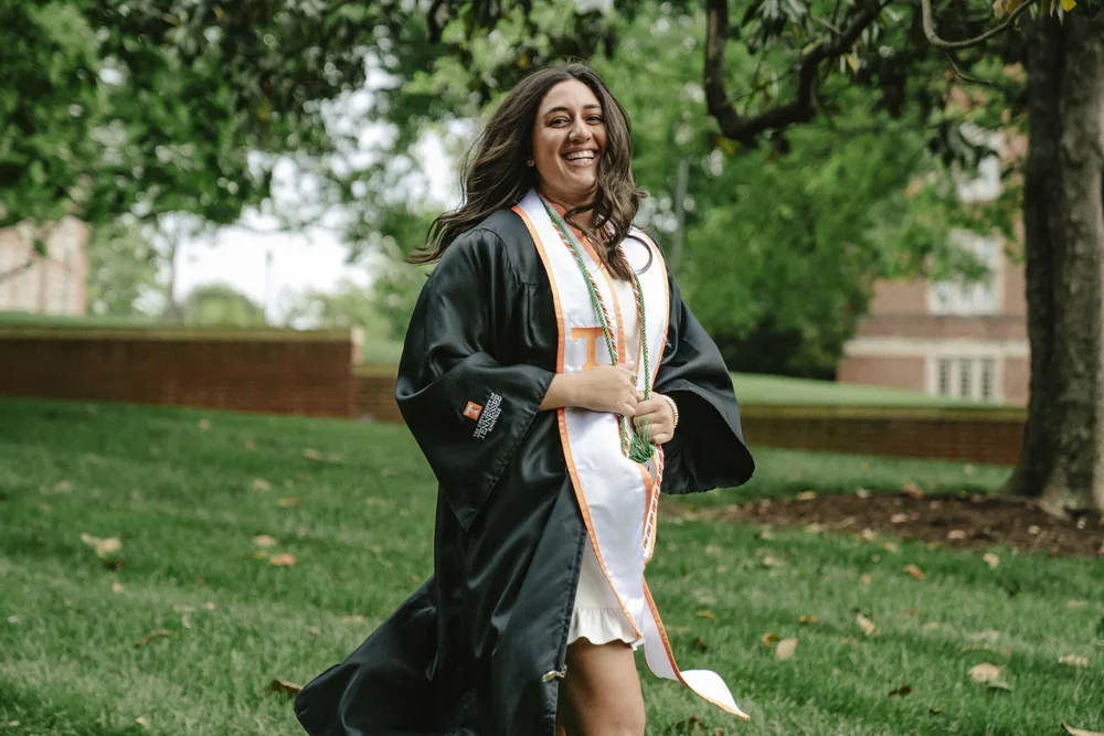 A woman in a graduation gown and stole standing outdoors on grass, smiling.