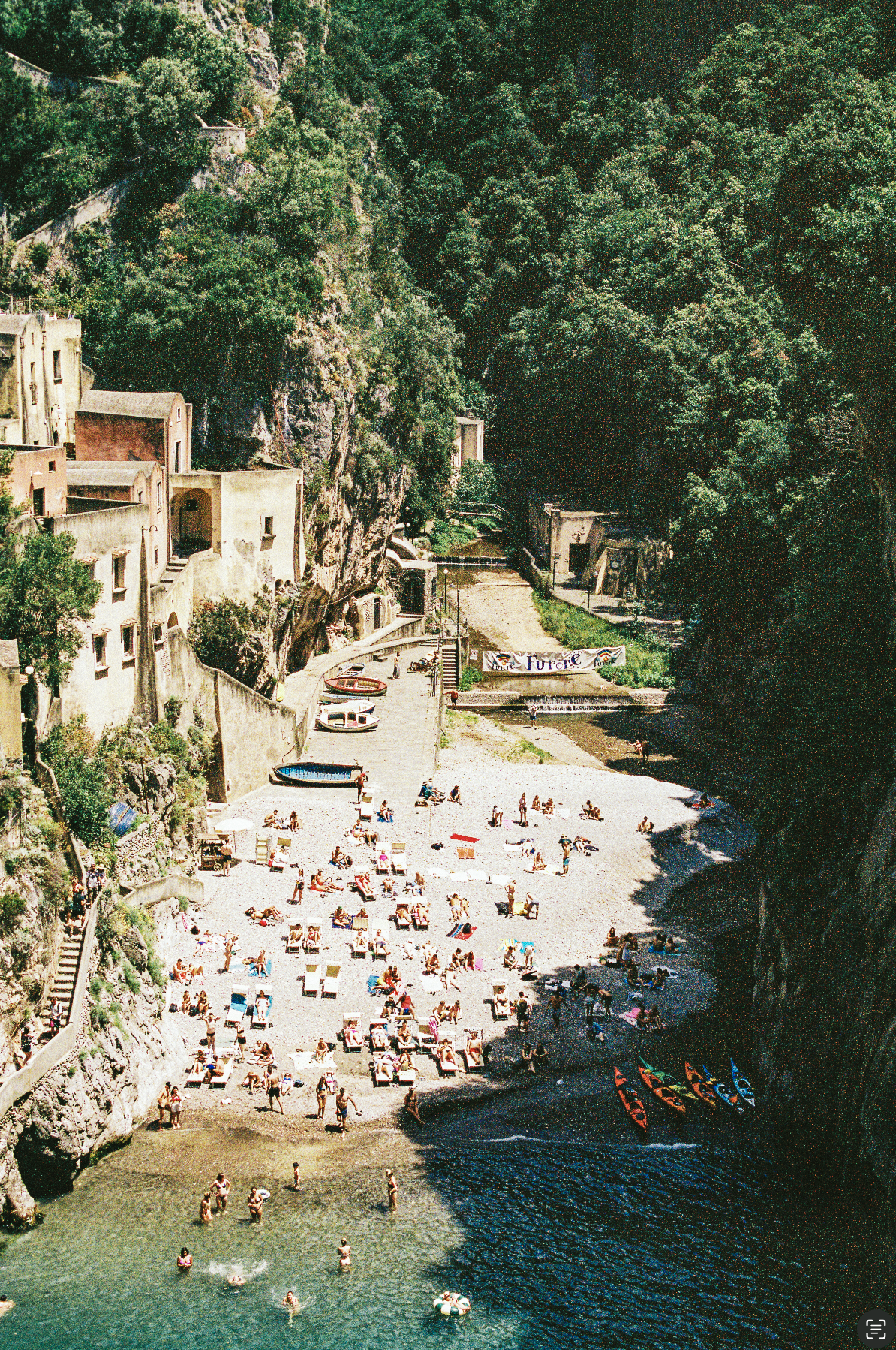 People relaxing on a beach with colorful umbrellas, boats, and kayaks, surrounded by green cliffs and buildings.