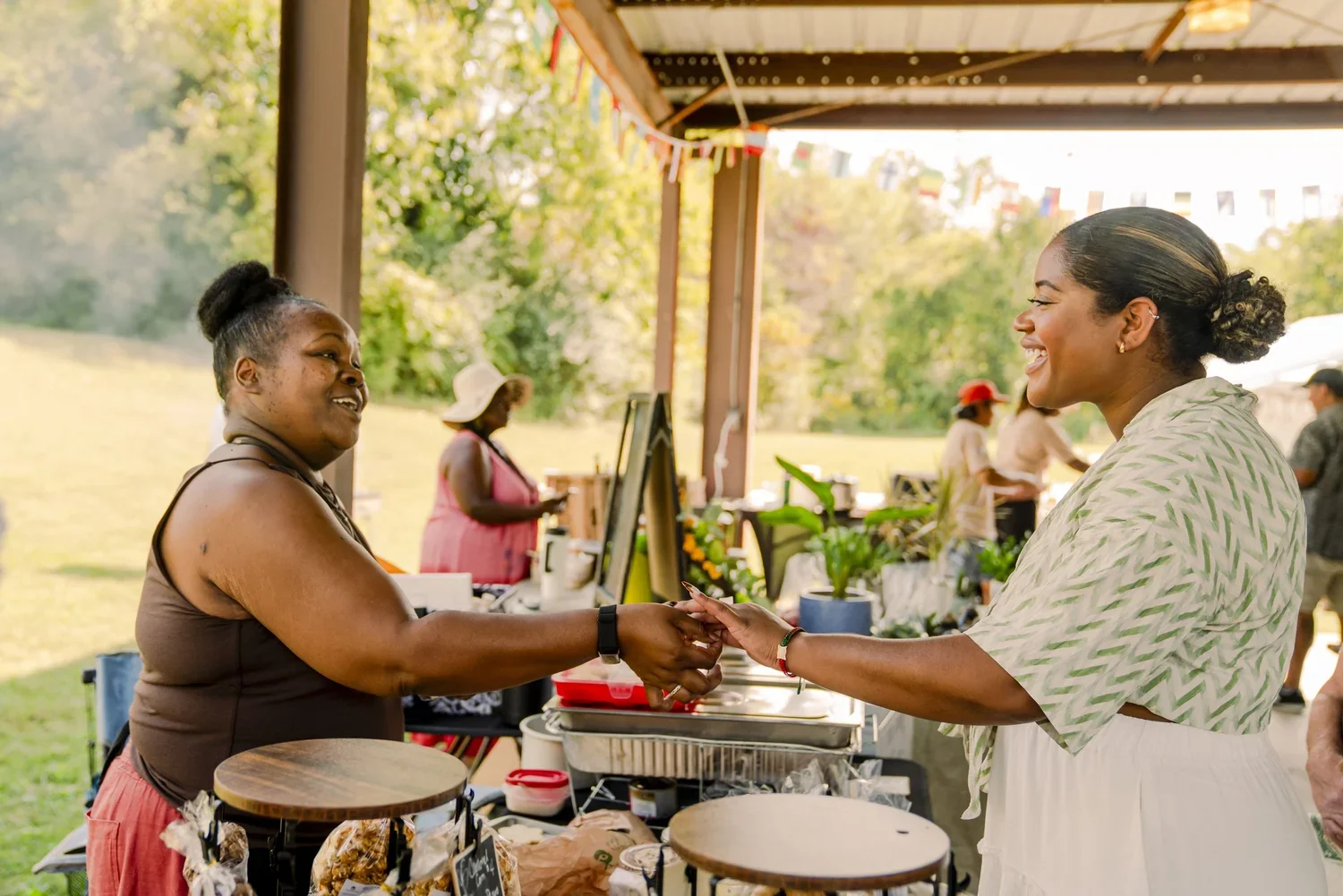 Two women smiling and holding hands at an outdoor market, with vendors and trees in the background.