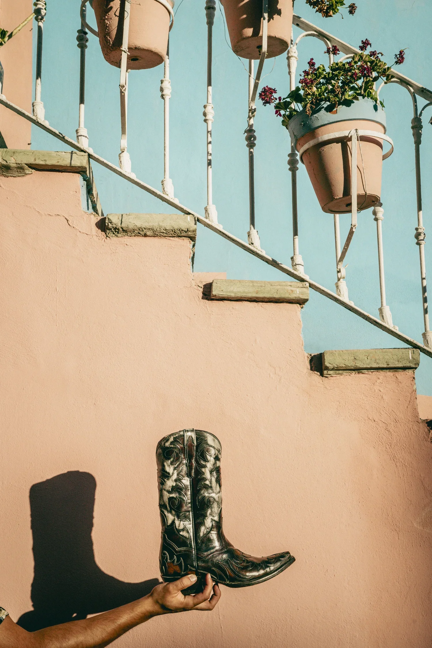 A person with a dark skin tone holding a black western cowboy boot with intricate white stitching, standing against a pink wall with a shadow cast on it. In the background, there is a staircase with potted plants and a bright blue sky.