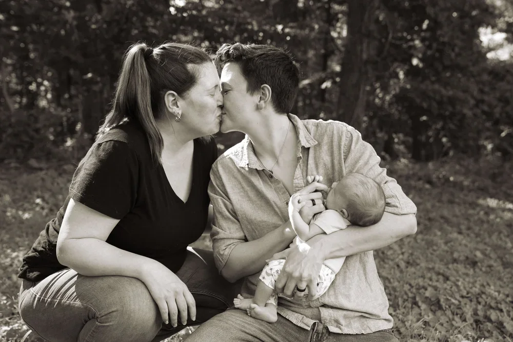 A woman and a person kiss while holding a baby outdoors, with trees in the background, in black and white.