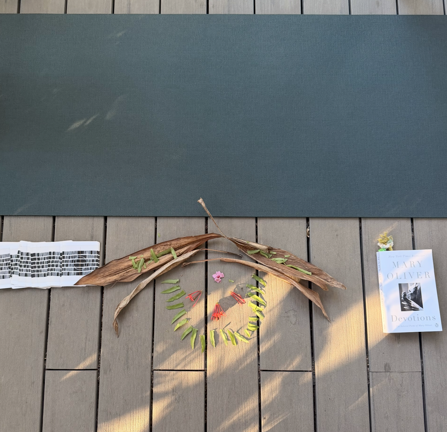 A yoga mat, a small stack of papers or cards, a mary oliver book titled 'Devotions,' and a circular arrangement of green leaves interwoven with dried brown leaves and red and pink flowers on a wooden deck.