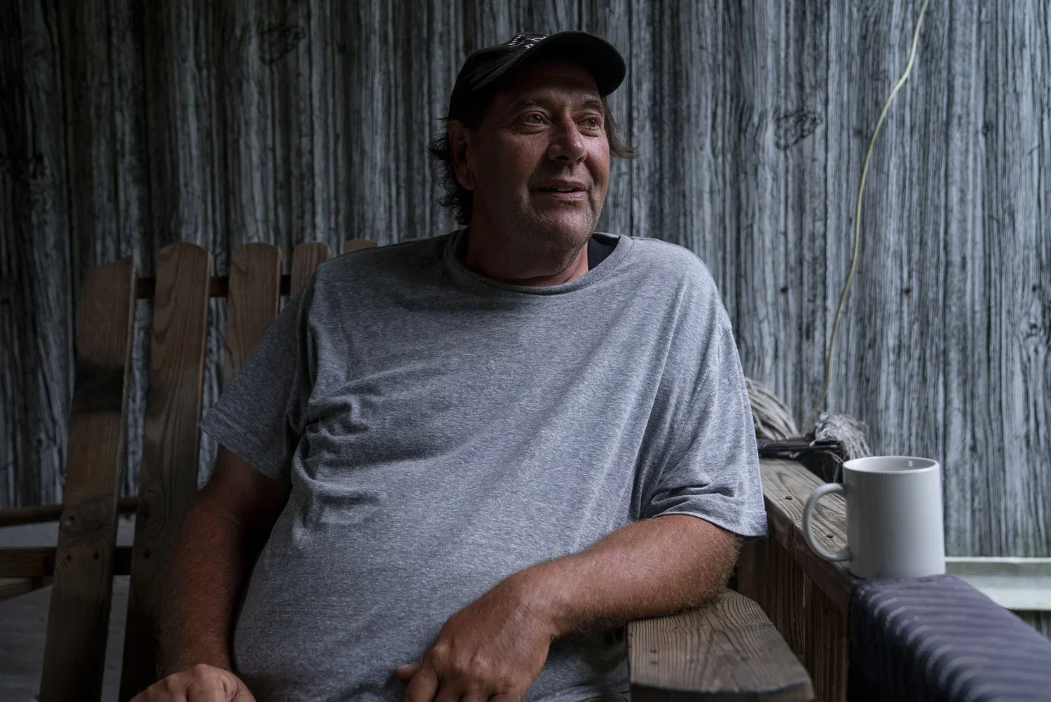 A middle-aged man in a gray t-shirt and black cap sitting on a wooden outdoor deck, looking to the side with a slight smile. A white mug is on the railing beside him, with a rustic wooden background behind him.