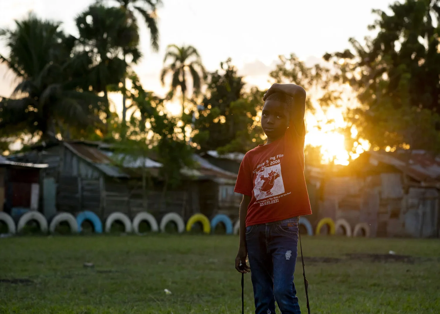 A young boy standing outdoors at sunset with his hand on his head, wearing a red t-shirt and jeans, with a row of tires painted in various colors in the background.