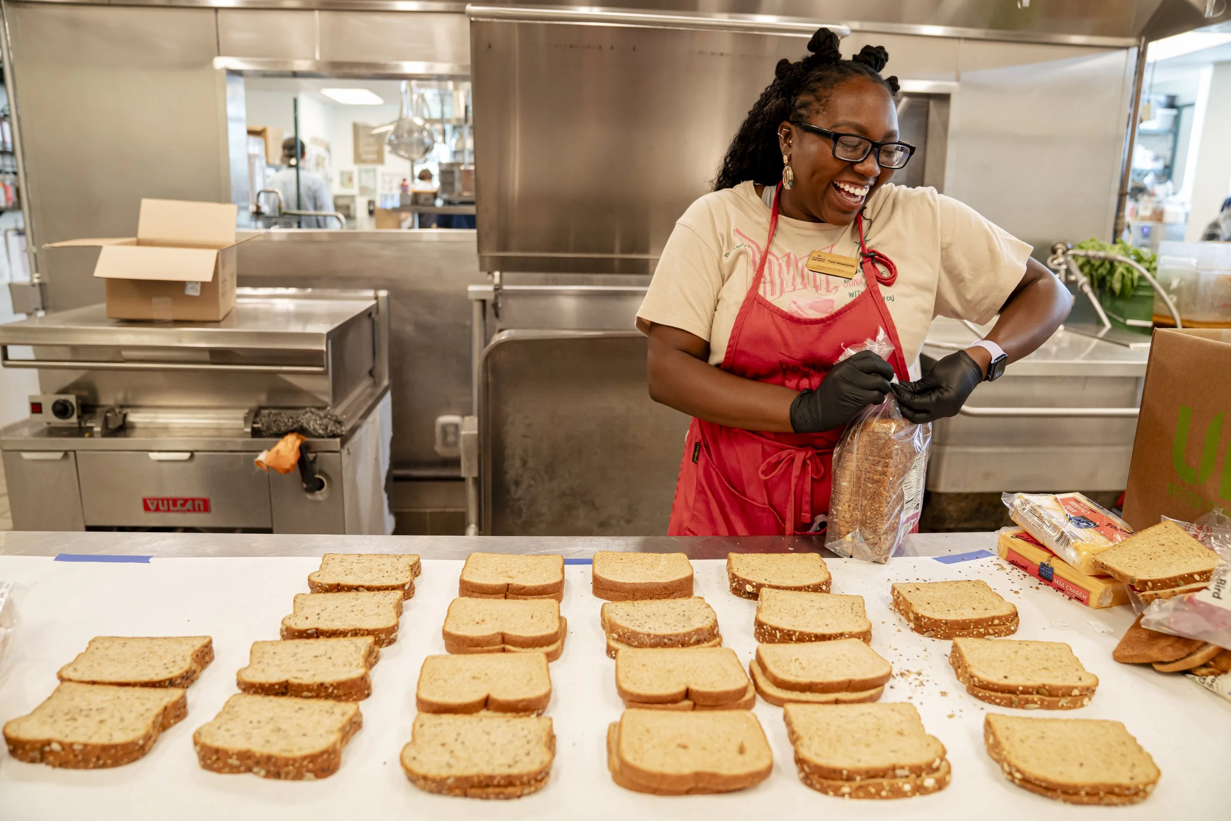 A woman wearing glasses, a beige T-shirt, a red apron, and black gloves, smiling while preparing sandwiches in a commercial kitchen. There are slices of bread on a white counter, some with spread, and a cardboard box and groceries nearby.
