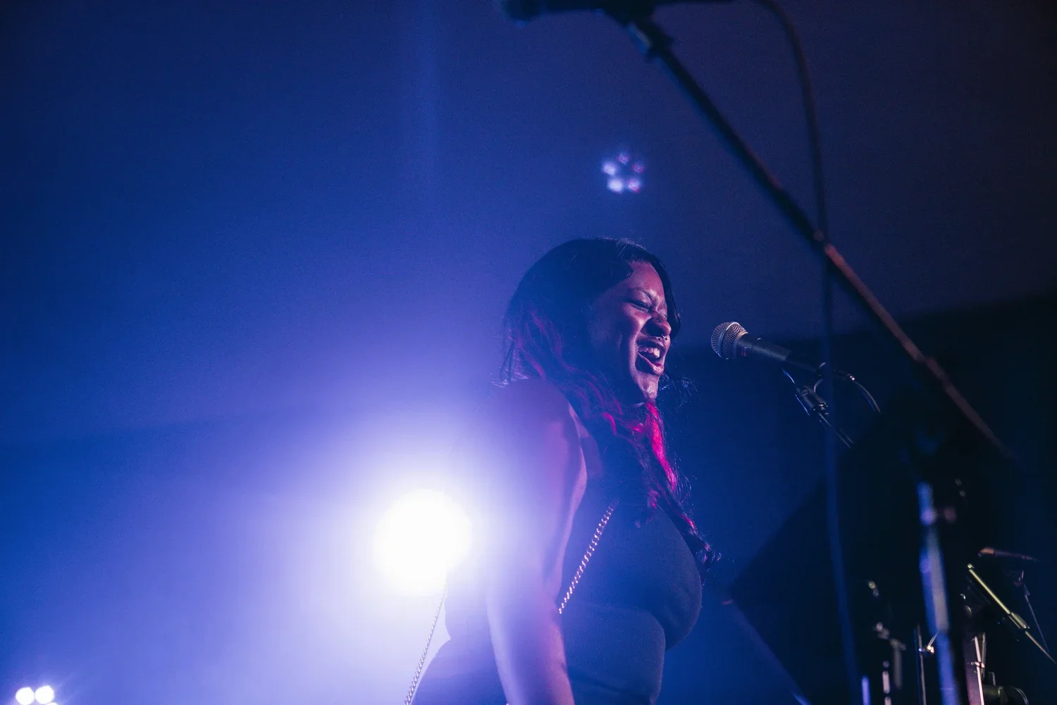 A woman singing passionately into a microphone on stage, backed by bright stage lights creating a glow around her.