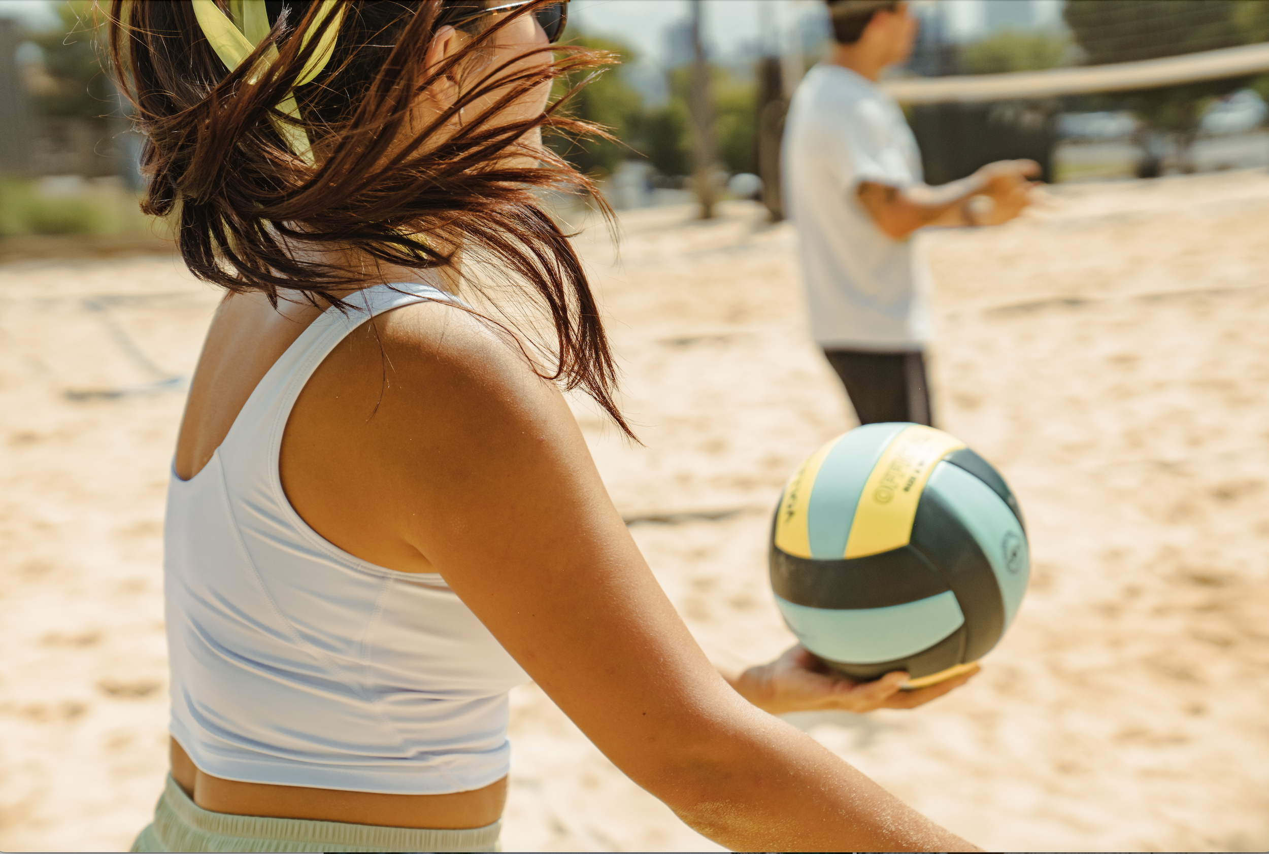 A woman with long, reddish-brown hair in a white tank top holding a volleyball at the beach, with a man in a white shirt and black shorts in the background.