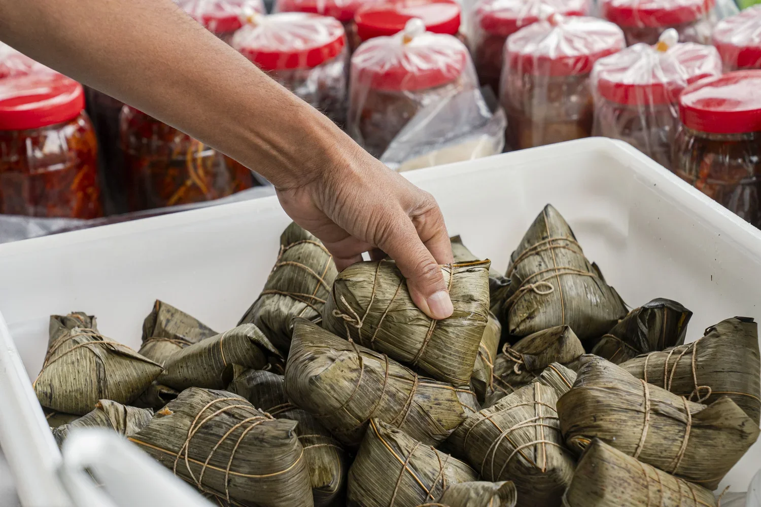 A person’s hand holding a traditional Asian rice cake wrapped in banana leaves, on a white tray, with jars of preserved vegetables in the background.
